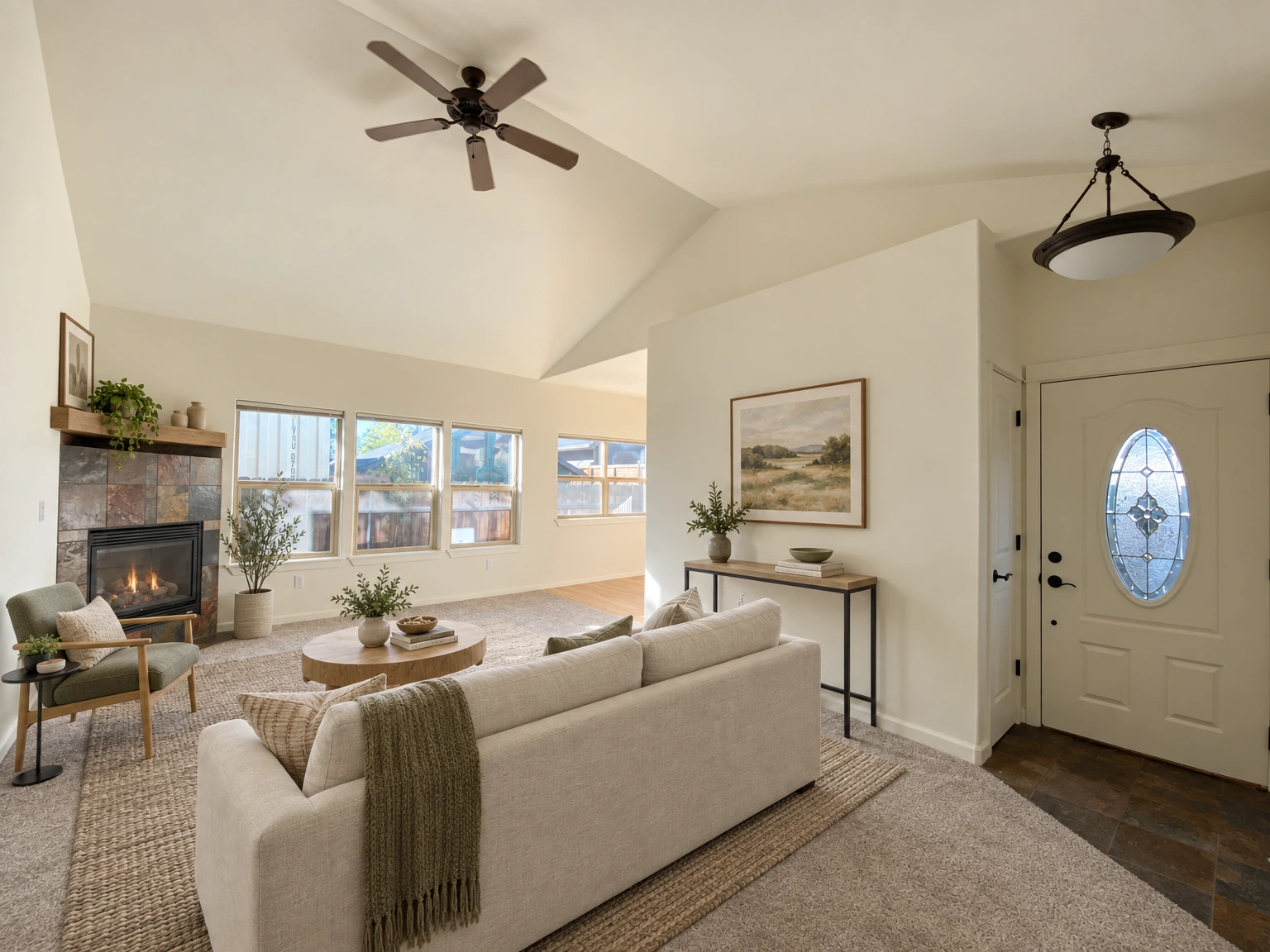 An empty carpeted room with a stone-faced fireplace, several windows, a ceiling fan, and a front door with an oval glass panel. (virtually staged)