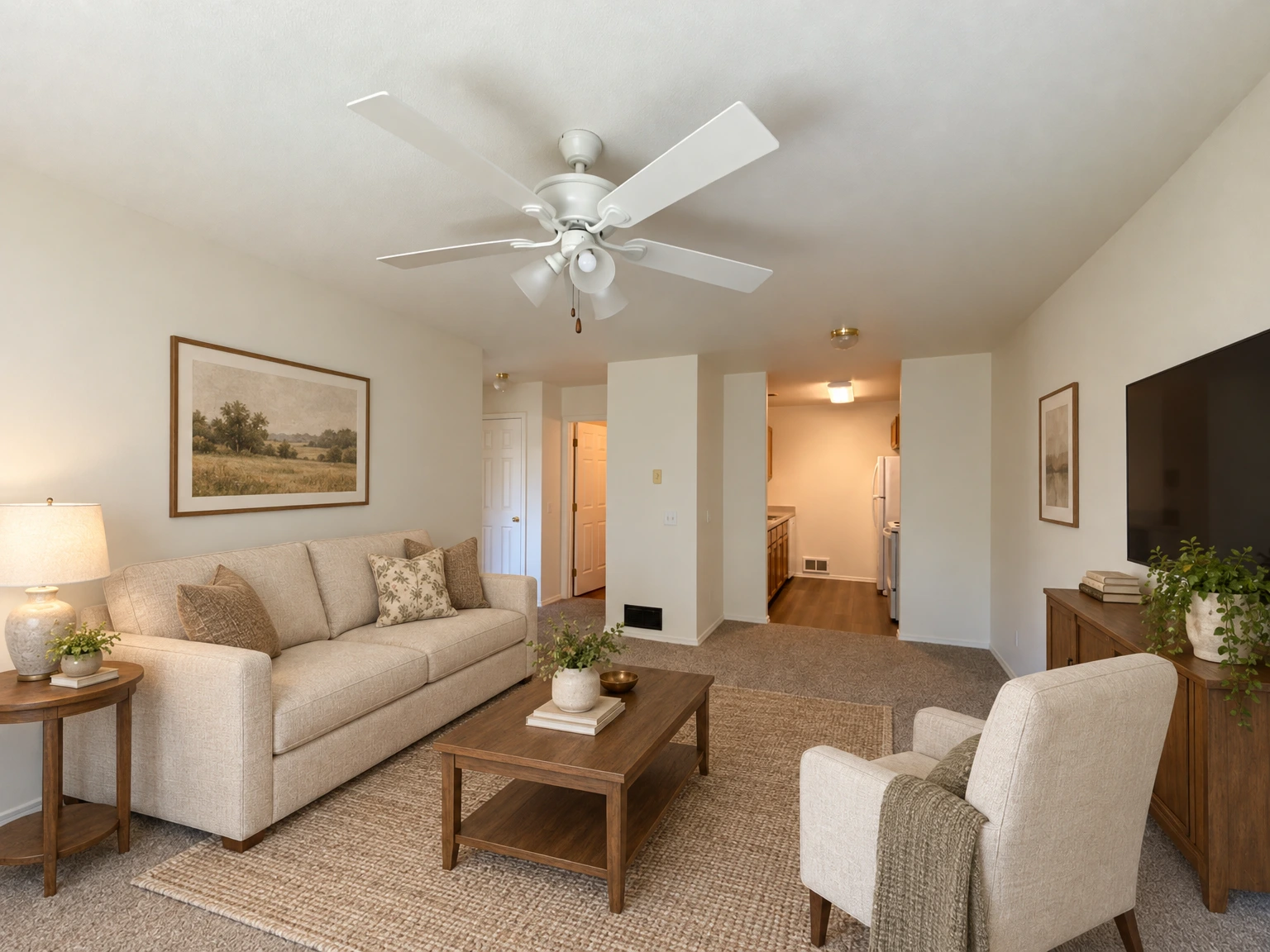 An empty carpeted room has white walls, a ceiling fan, and an opening to a kitchen with cabinets and a refrigerator. (virtually staged)