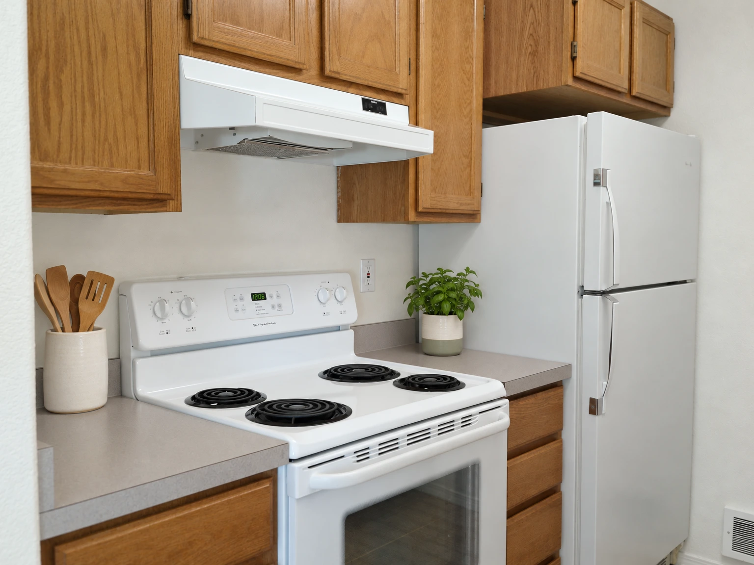 A white electric stove with four coil burners sits beneath a range hood, beside a white refrigerator and wooden cabinets. (virtually staged)