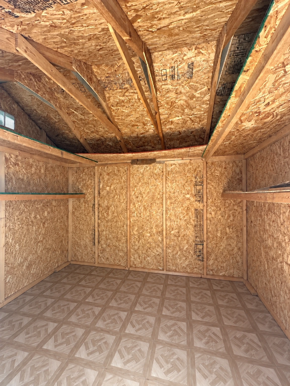 An unfinished wooden room has exposed framing and sheathing, a patterned floor, and two panels leaning against the left wall. (virtually staged)