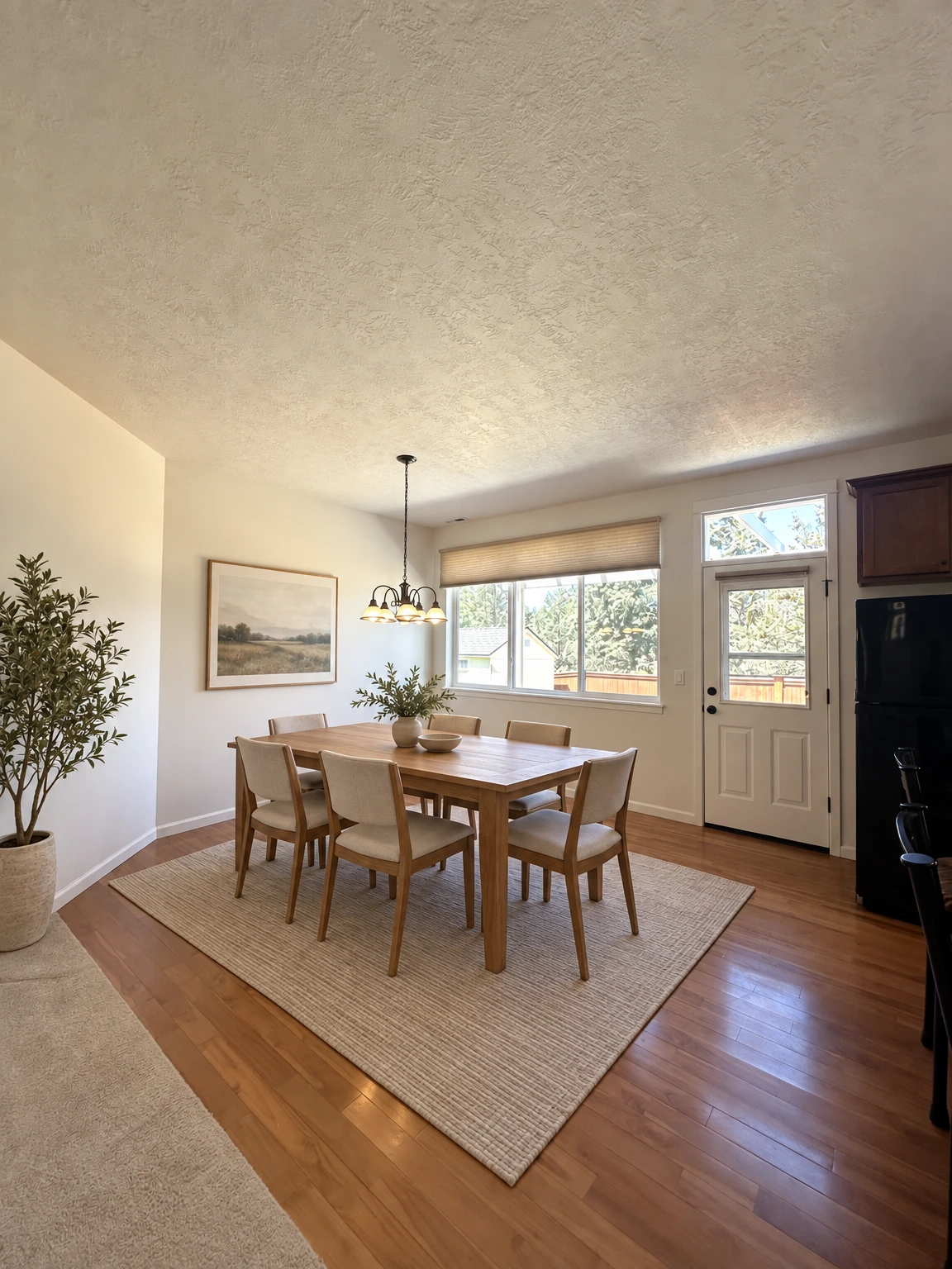 An empty room has wood flooring, a textured ceiling, a hanging light, a window with a shade, and a glass-paneled door. (virtually staged)