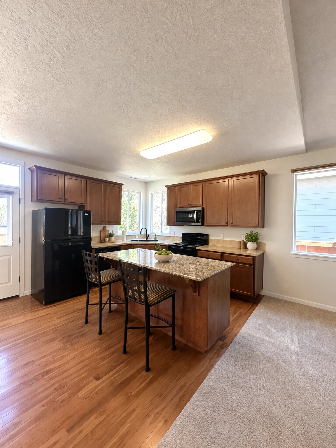 A kitchen with dark wood cabinets, a granite-topped island and chairs, black appliances, and windows above the sink. (virtually staged)