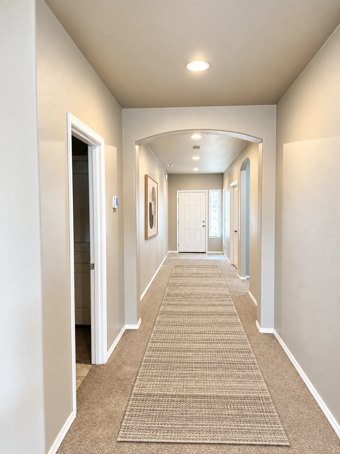 A carpeted hallway with beige walls, an arched opening, several doorways, and a white door at the far end. (virtually staged)