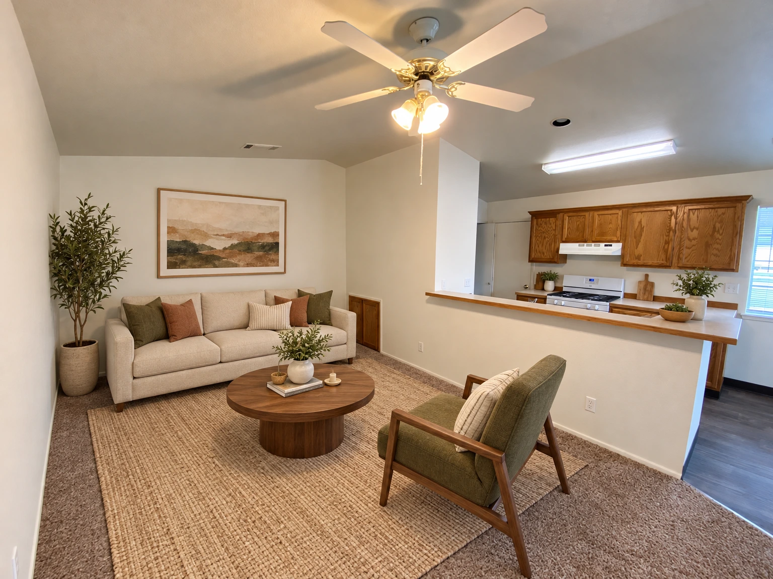 An empty carpeted room opens to a kitchen with wood cabinets and a stove; a ceiling fan with lights hangs overhead. (virtually staged)