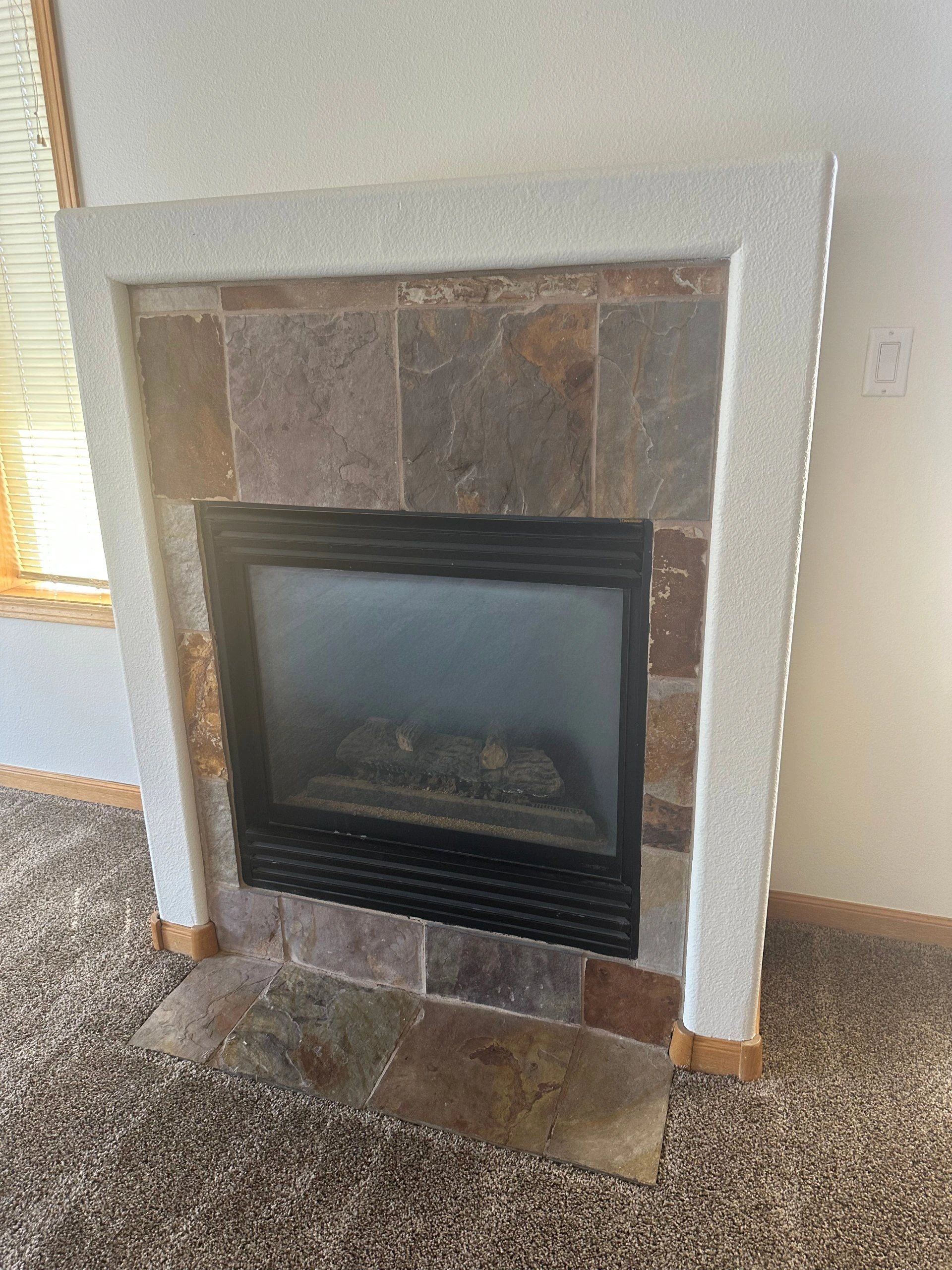 A black glass-fronted fireplace is set in a multicolored stone surround beneath a wide white mantel.