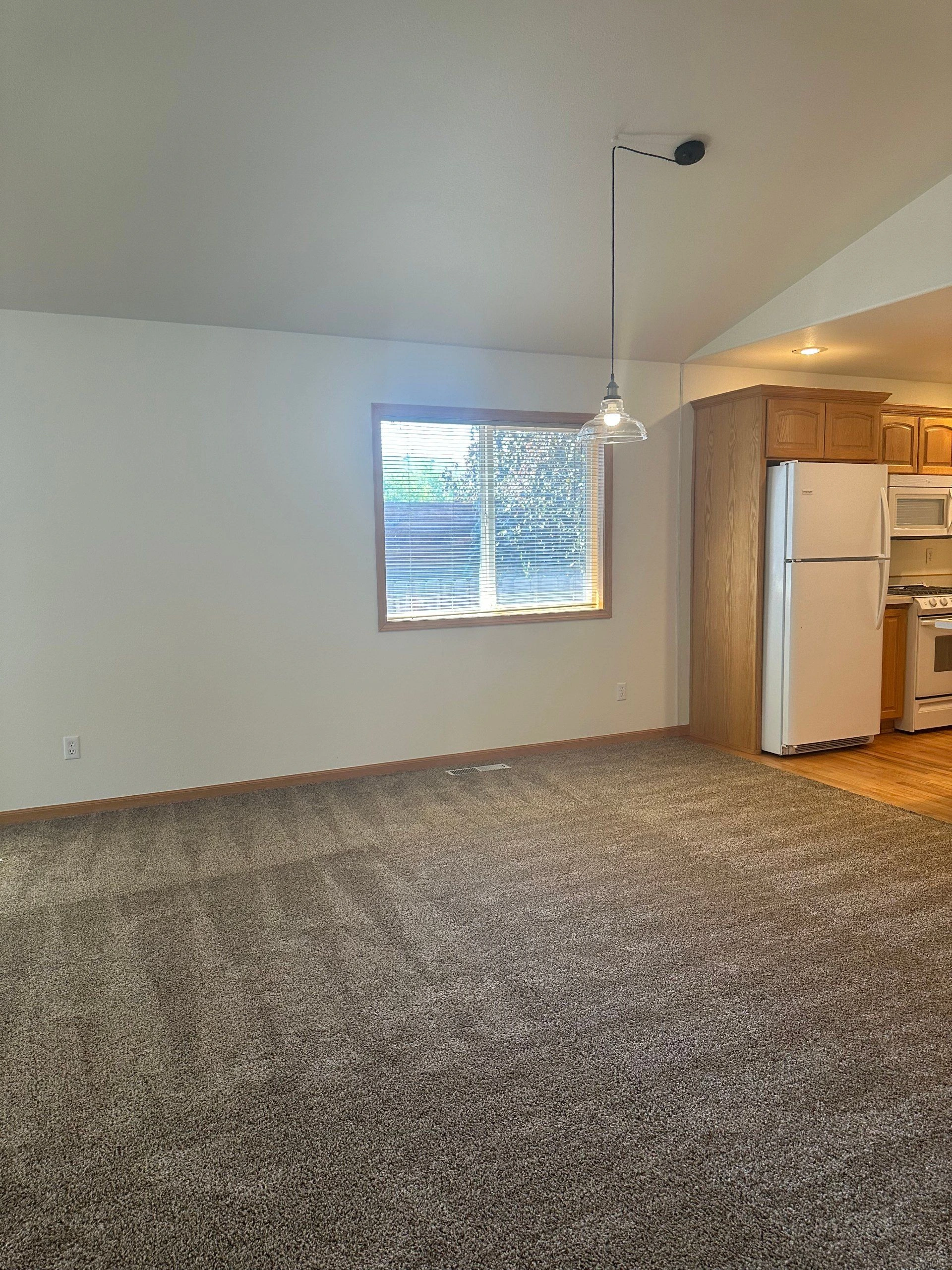 A carpeted room opens to a kitchen with wood cabinets; a window with blinds and a hanging glass light are visible.
