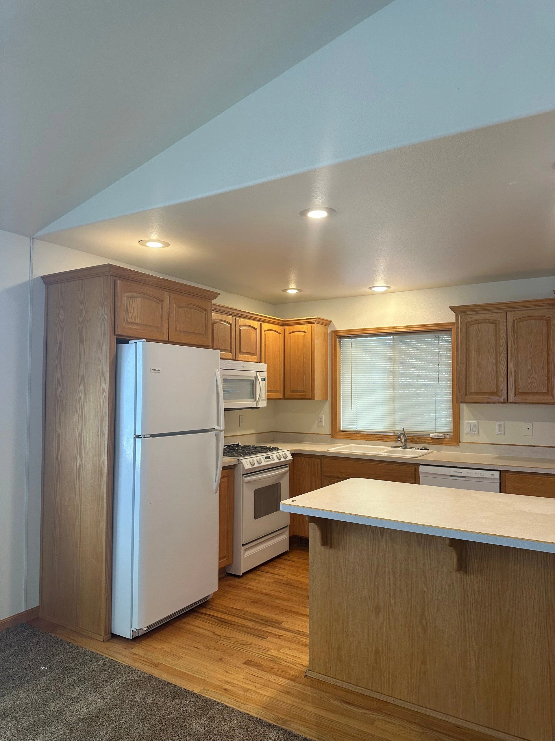 A kitchen with wood cabinets, white appliances, a sink beneath a window with blinds, and a large island.