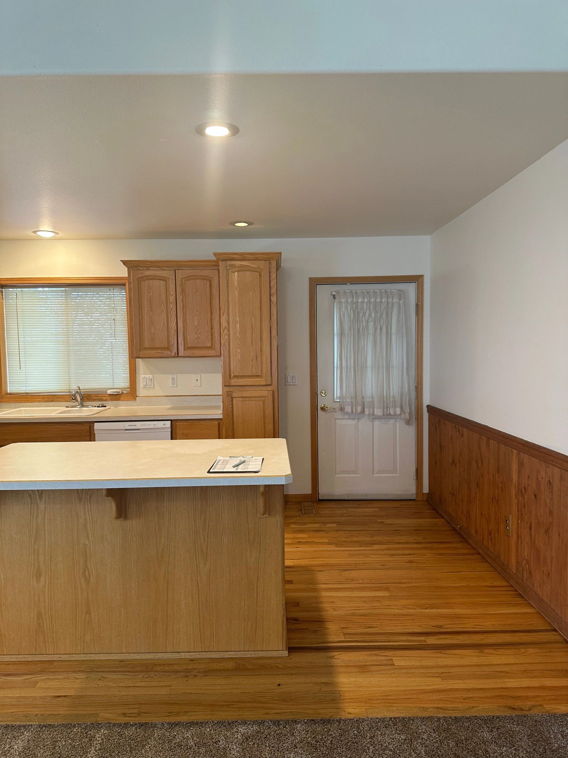 A kitchen with wood cabinets, a sink beneath a window, a large island, and a curtained door.