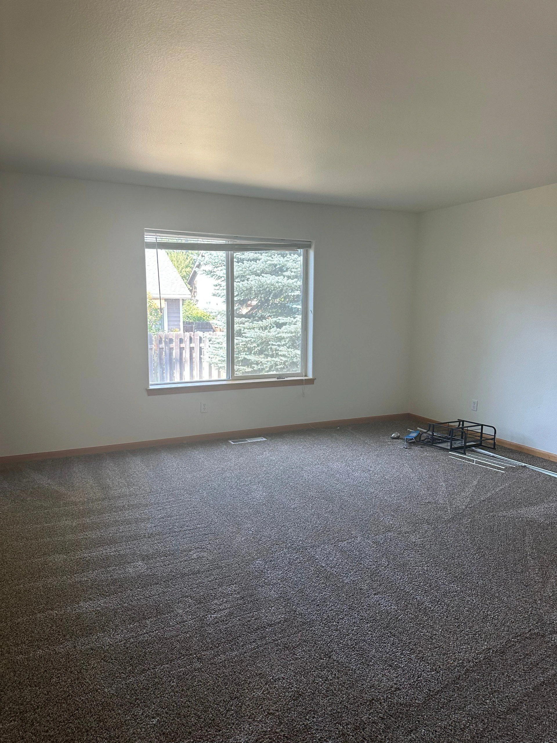 An empty room with gray carpet, pale walls, a window with blinds, and metal frame pieces on the floor.