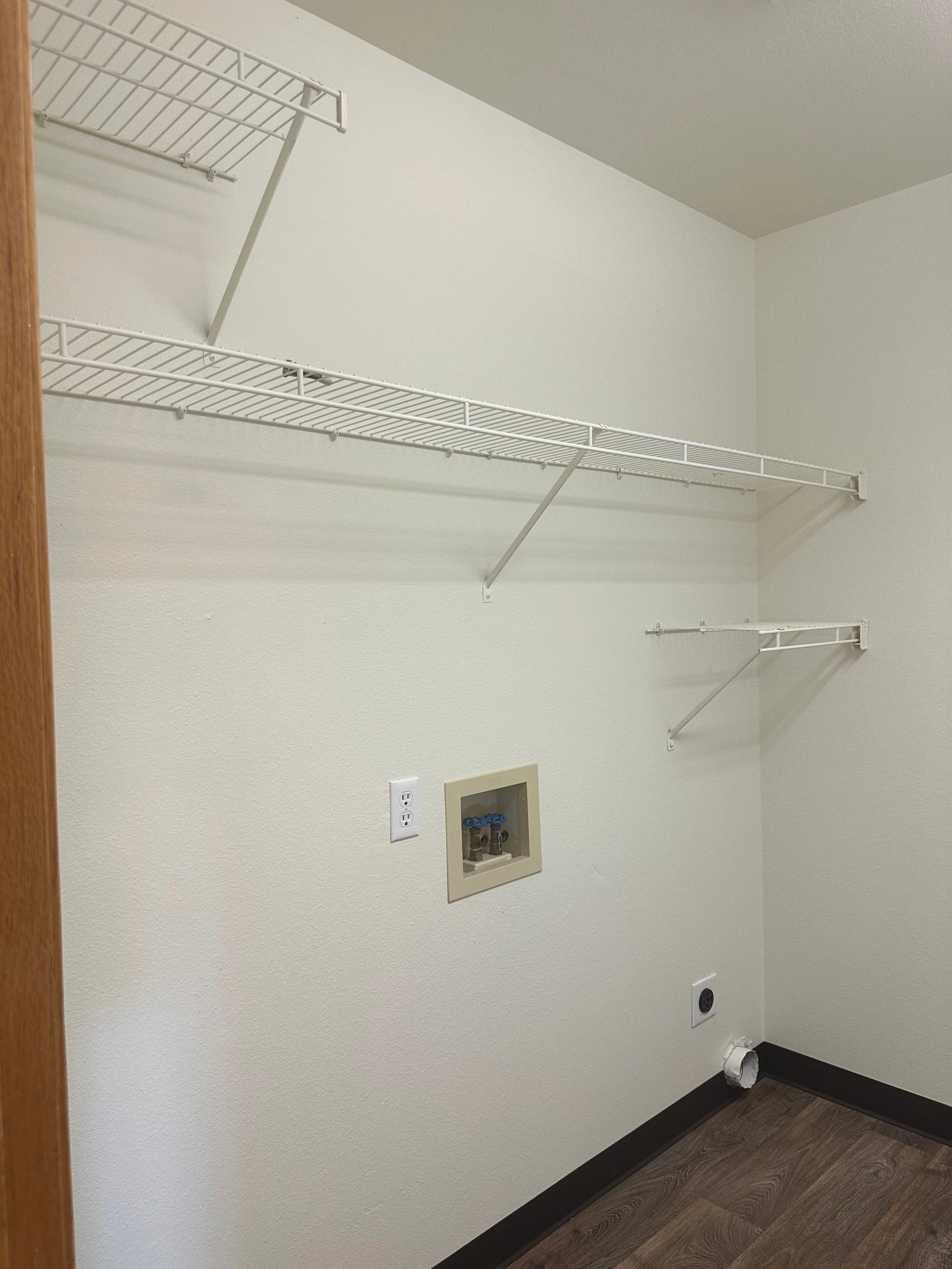 A white-walled space has several wire shelves, wall hookups and outlets, with dark wood-pattern flooring.