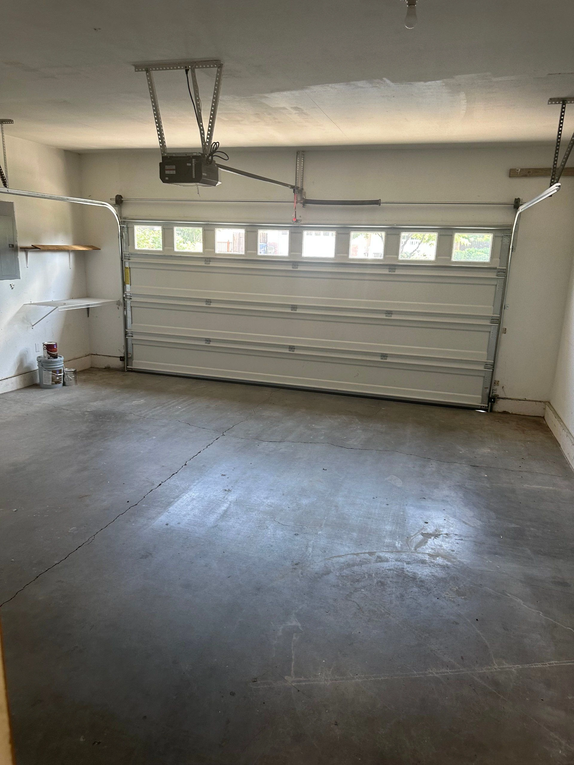A garage with a closed sectional door, a row of windows, an overhead opener, and a cracked concrete floor.