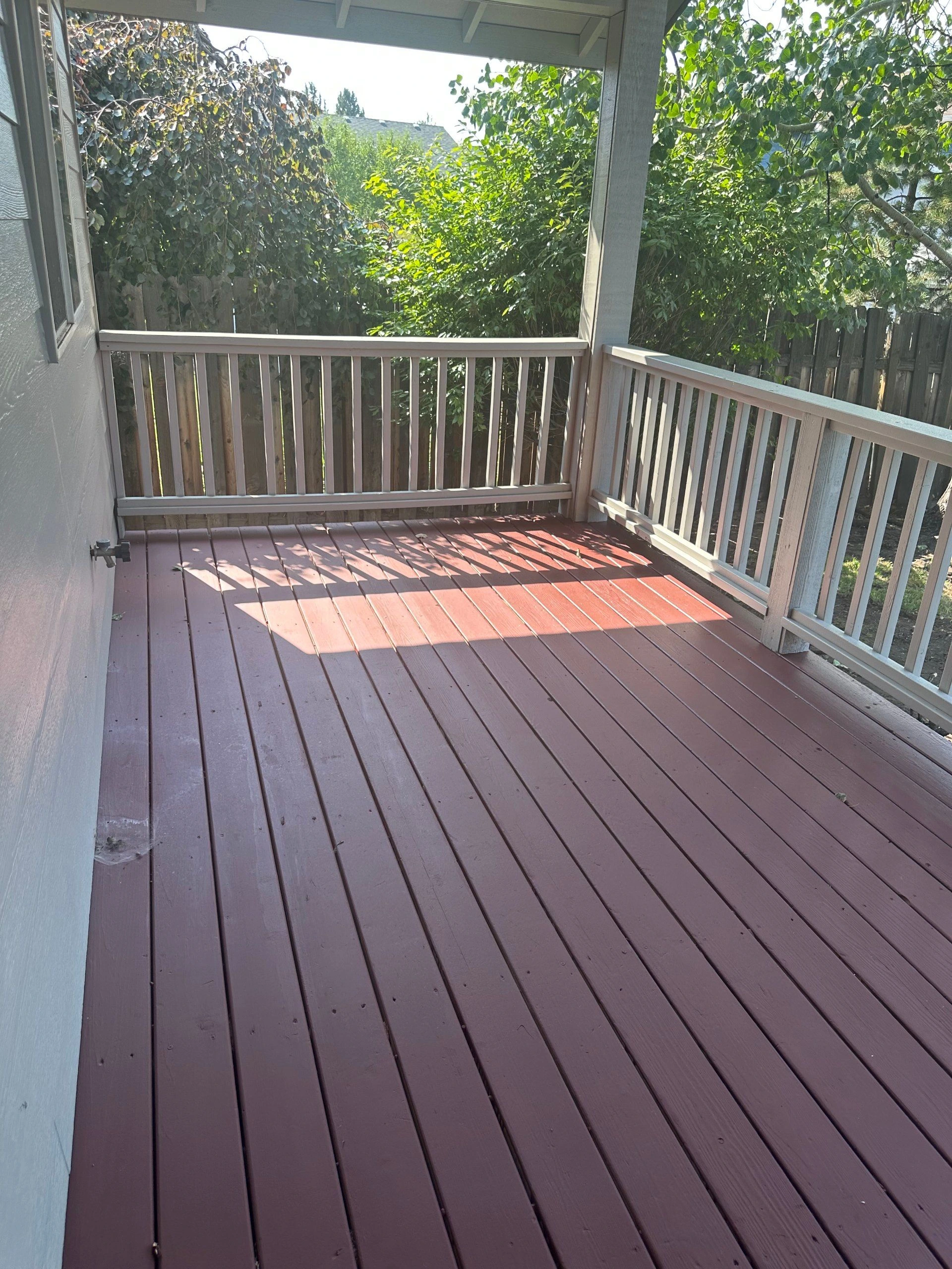A covered deck with reddish plank flooring and pale railings, surrounded by leafy trees and shrubs.