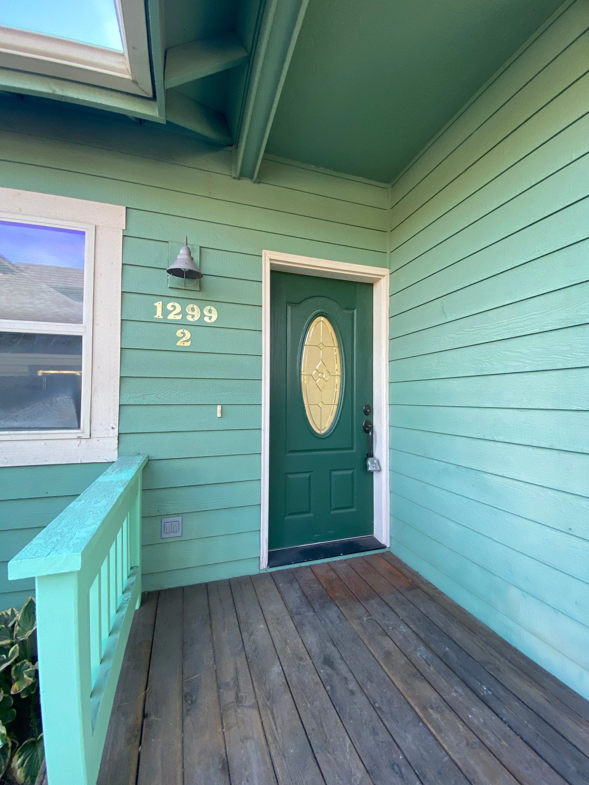 A covered porch has a dark green door with an oval glass panel, turquoise siding, a window, and a wall lamp.