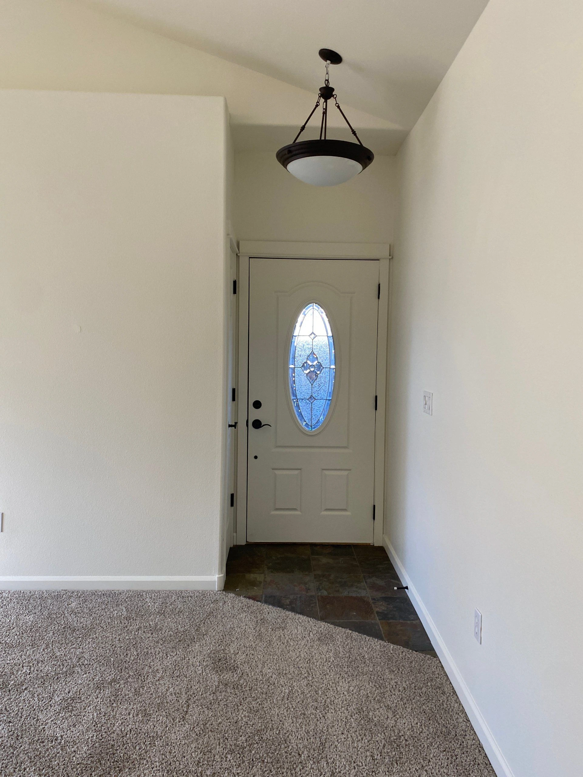 A white entry door with an oval blue glass panel stands beneath a hanging light, beside carpet and dark tile flooring.
