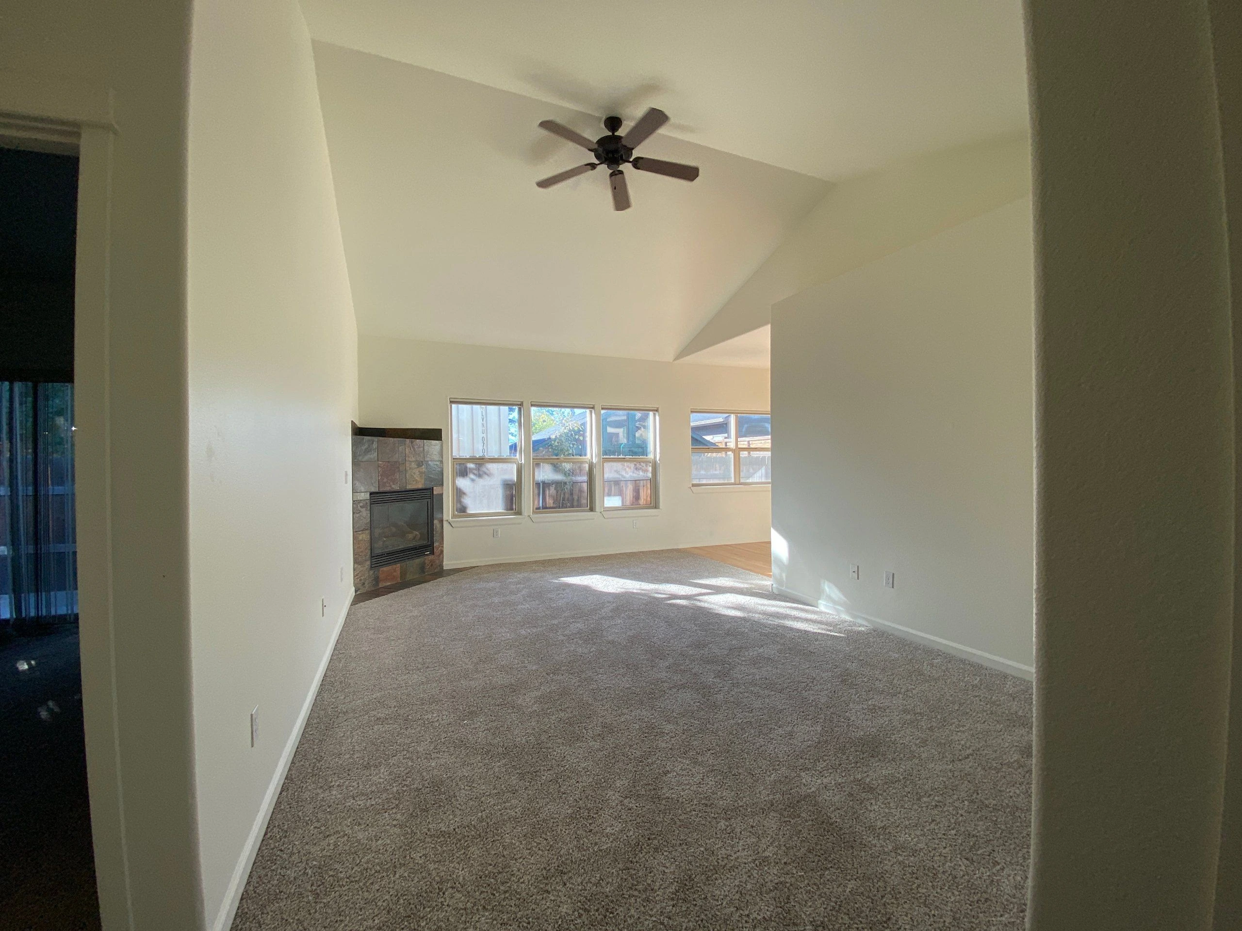 An empty carpeted room has a high angled ceiling, a ceiling fan, a tiled fireplace, and several windows.