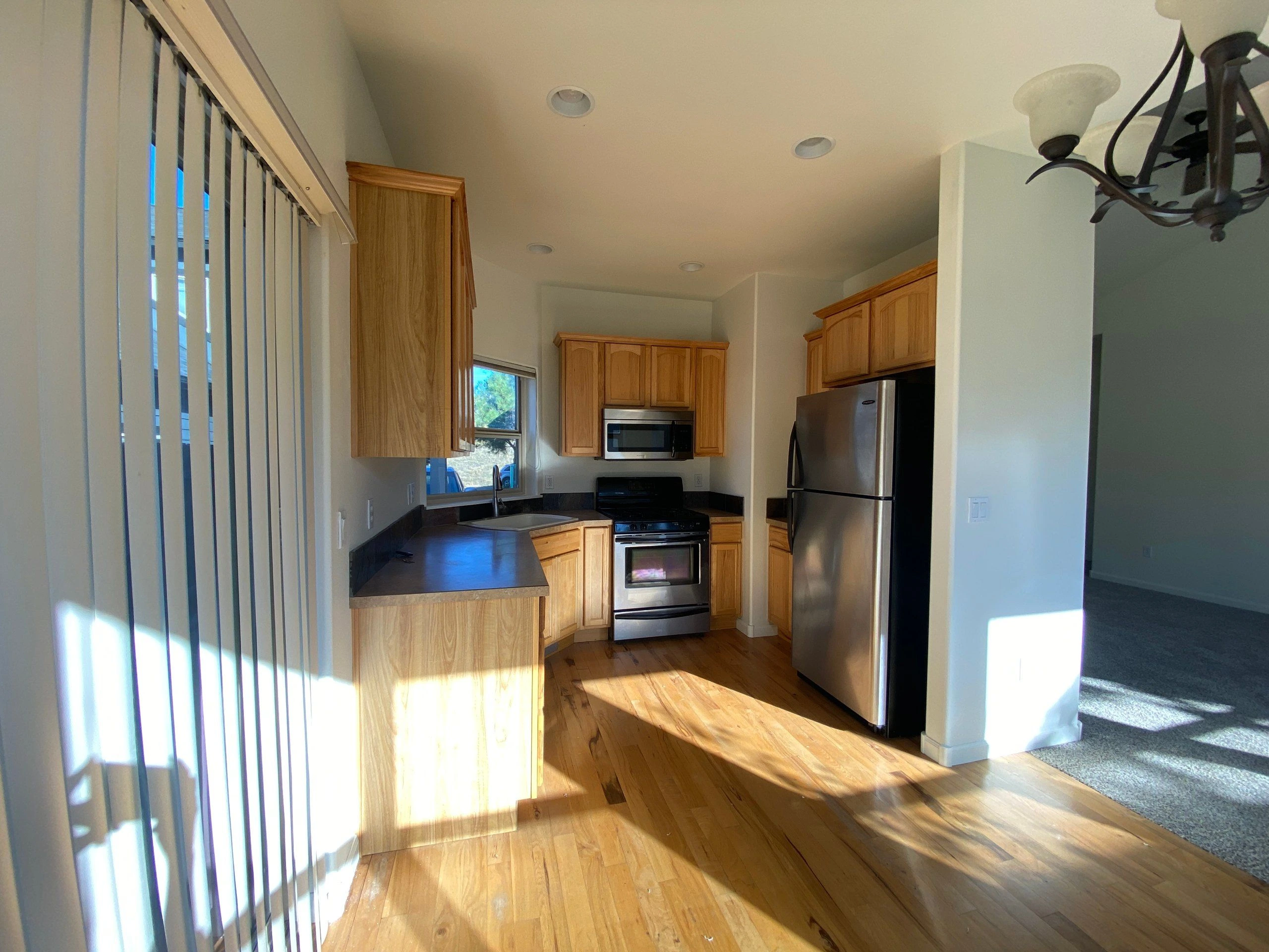 A kitchen with wood cabinets, dark counters, a stainless-steel refrigerator, a range and microwave, and a sink beneath a window.