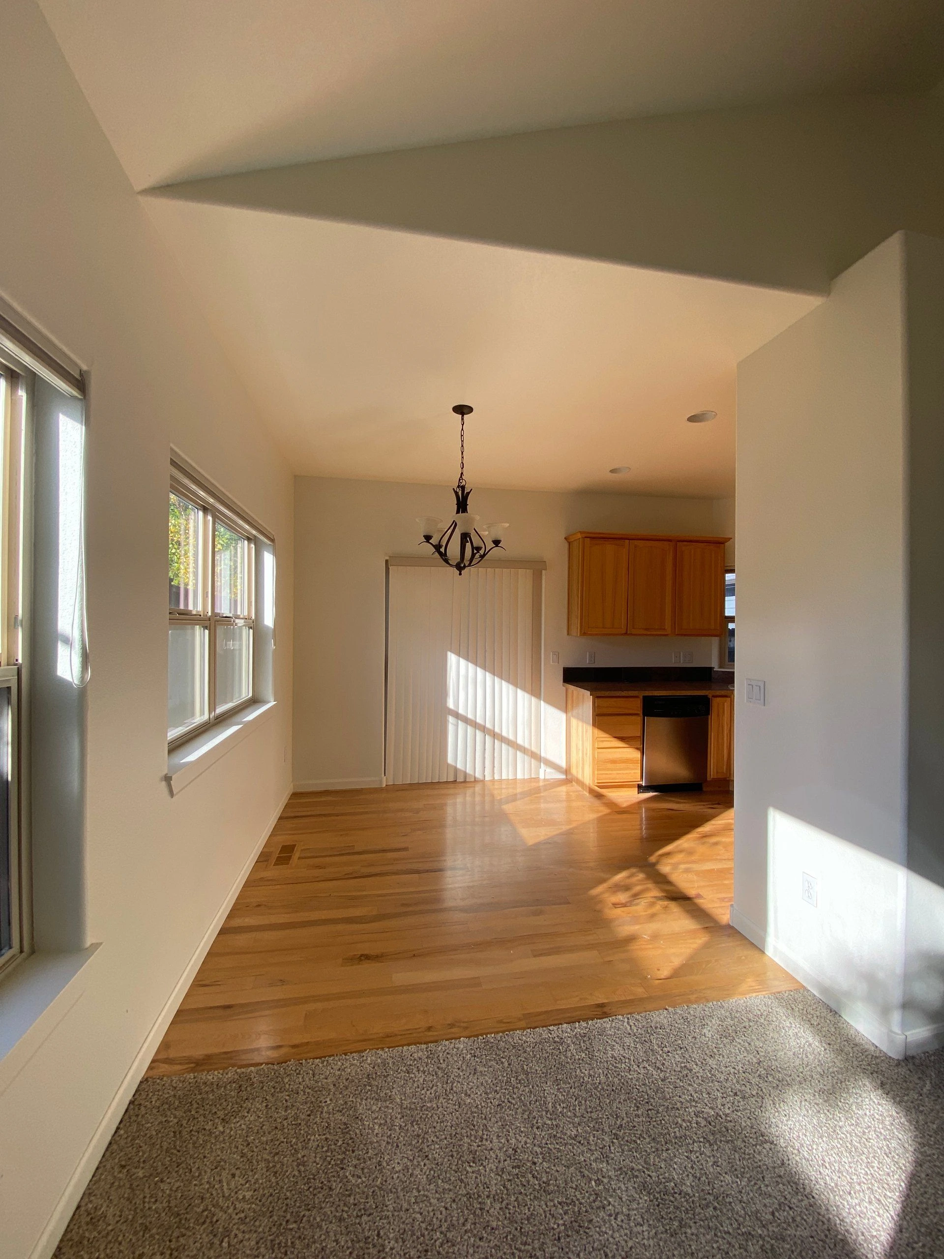 A sunlit room has large windows, wood flooring, a carpeted foreground, a hanging chandelier, and a kitchen with wood cabinets.