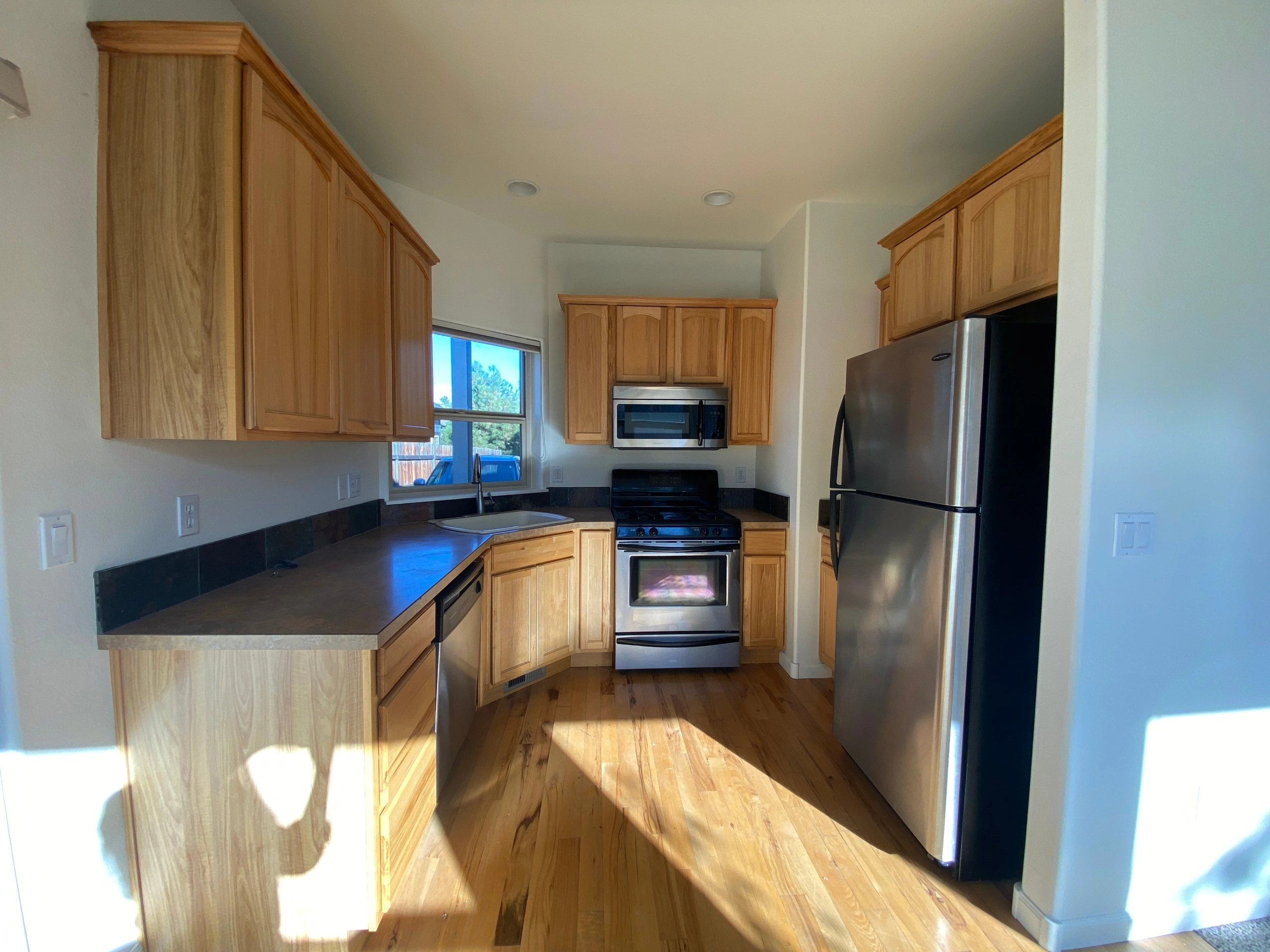 A kitchen with wooden cabinets, dark counters, a sink beneath a window, a stove, microwave, and stainless-steel refrigerator.