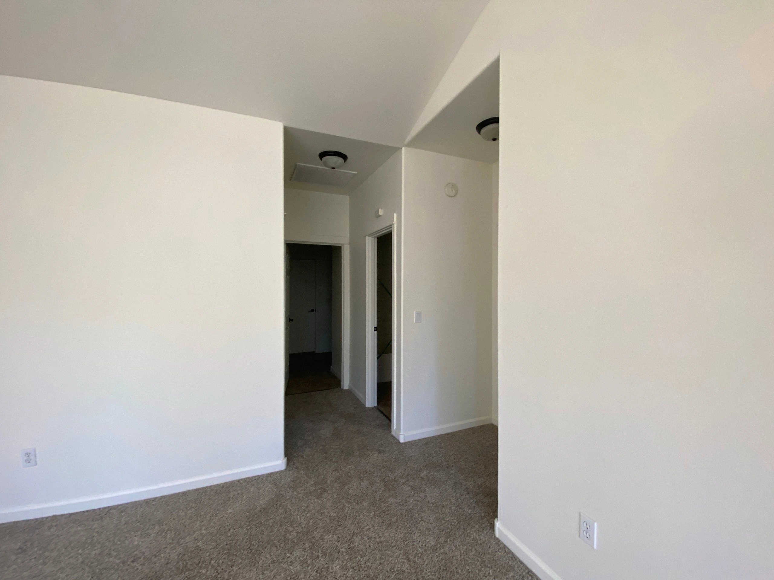 An empty carpeted room opens onto a hallway with two doorways and white walls.