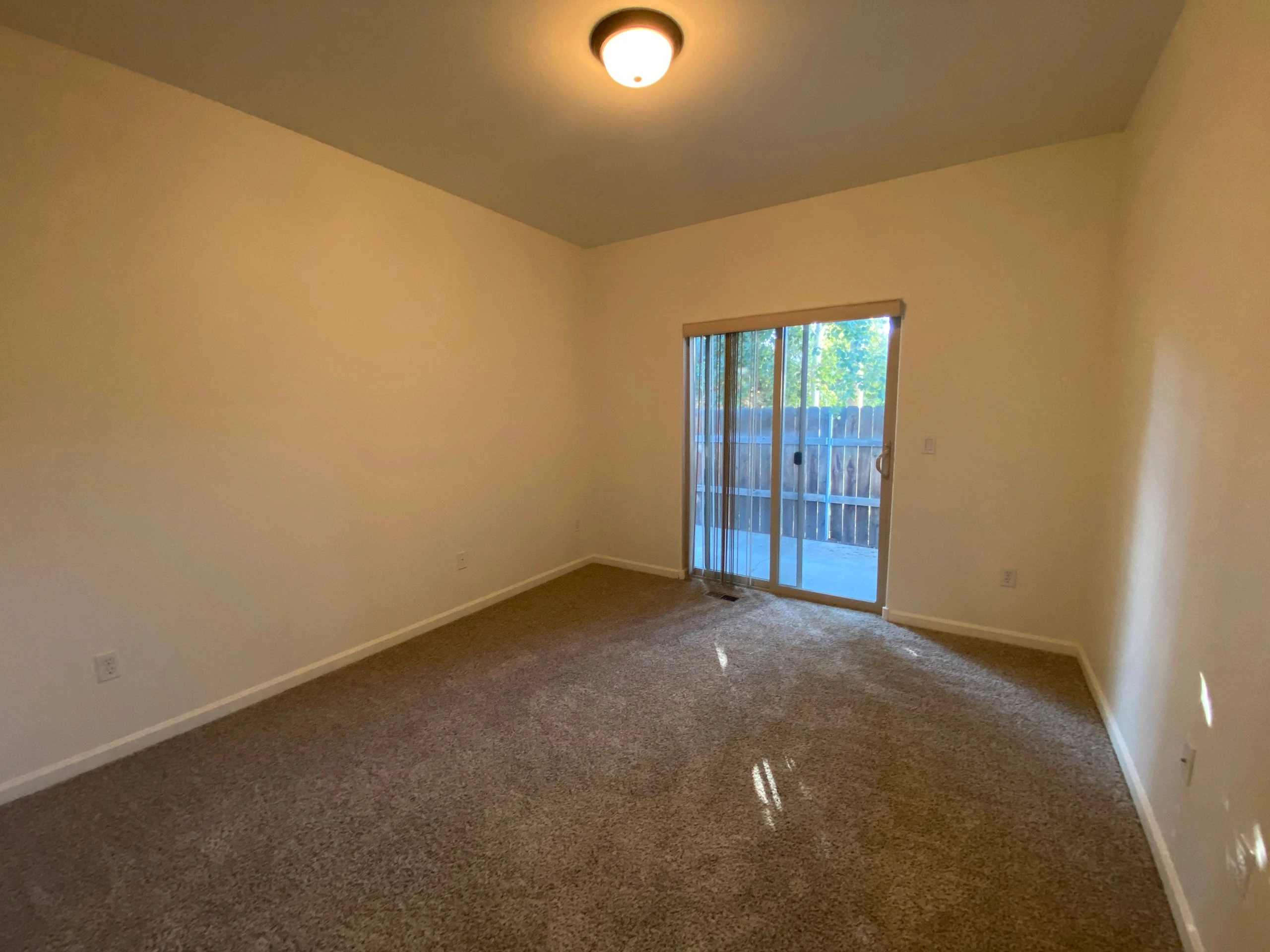 An empty room with beige walls, brown carpet, a ceiling light, and a sliding glass door with vertical blinds.