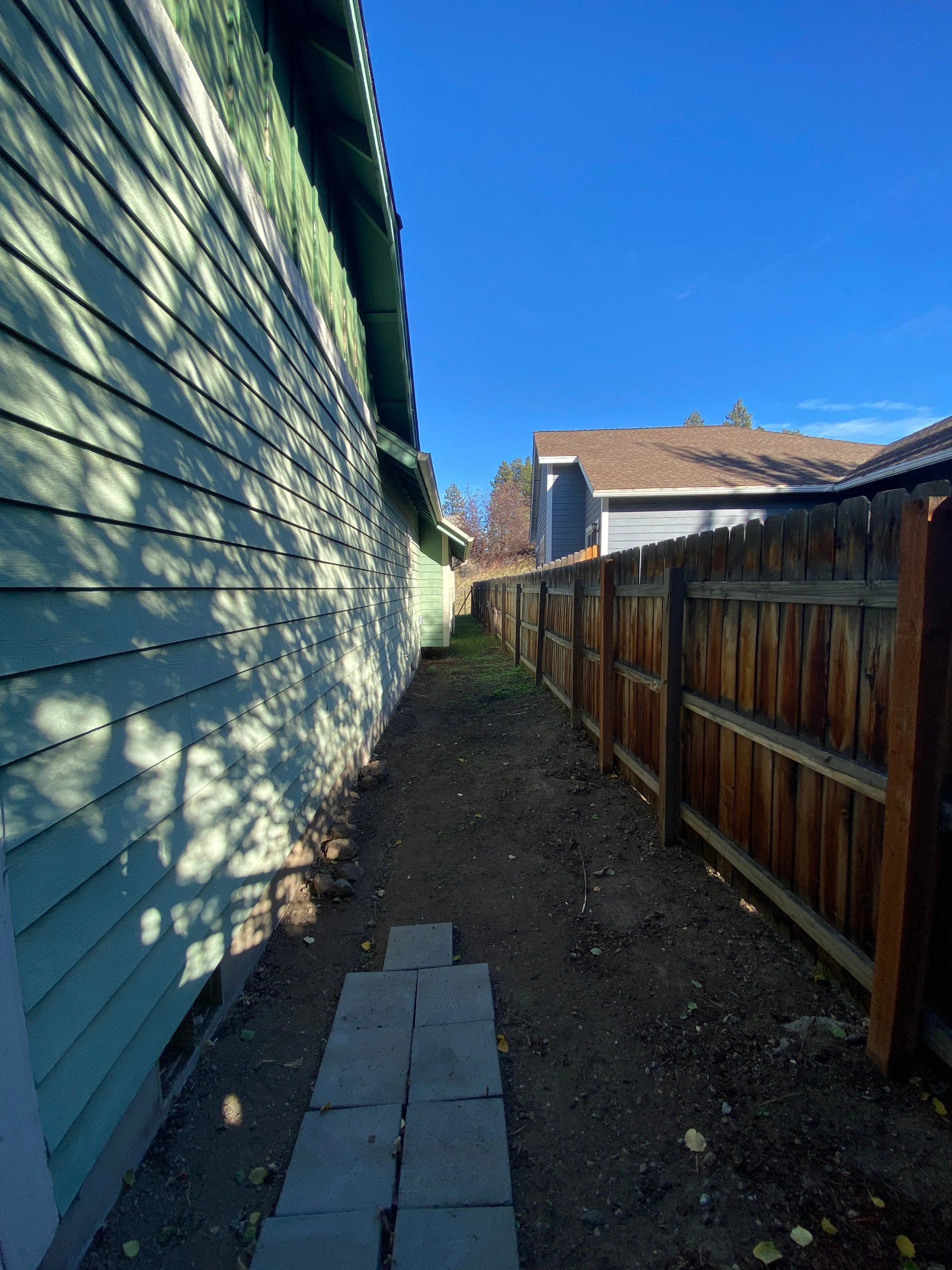 A narrow dirt path with stepping stones runs between a green-sided building and a wooden fence beneath a blue sky.