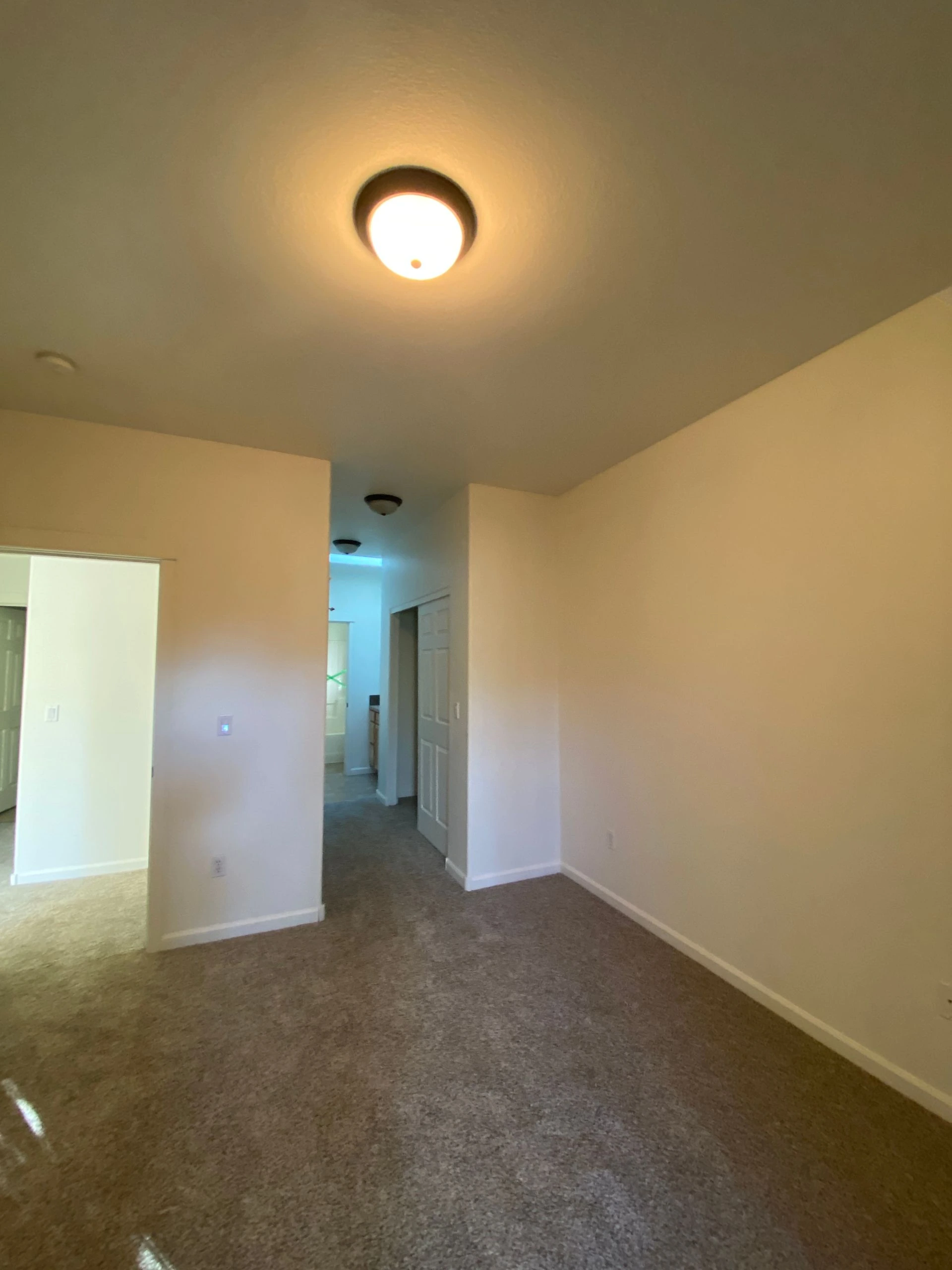 An empty carpeted room has beige walls, a lit ceiling fixture, and a hallway with doors at the back.