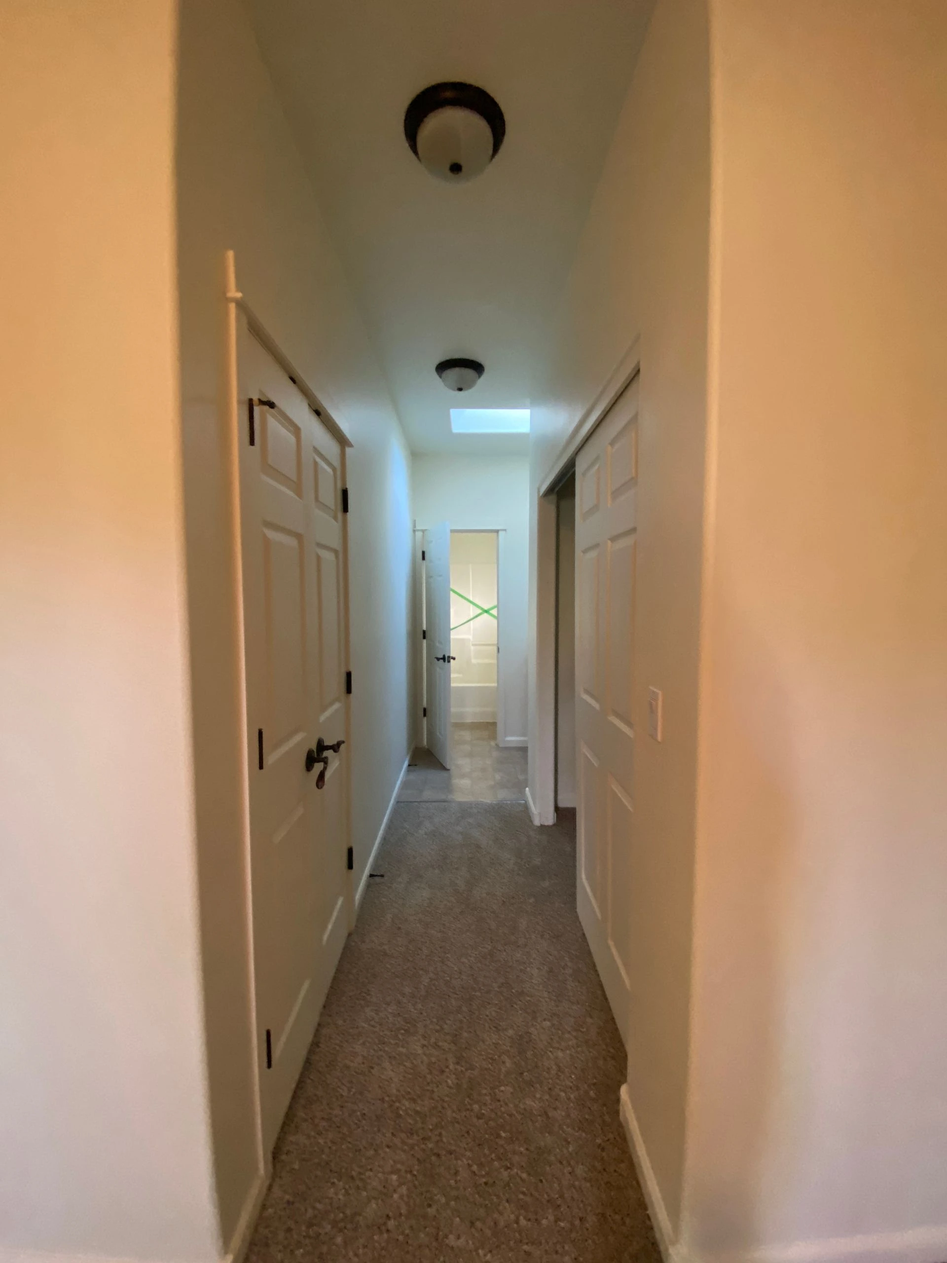 A narrow hallway with carpet, several paneled doors, two ceiling lights, and a rectangular skylight.