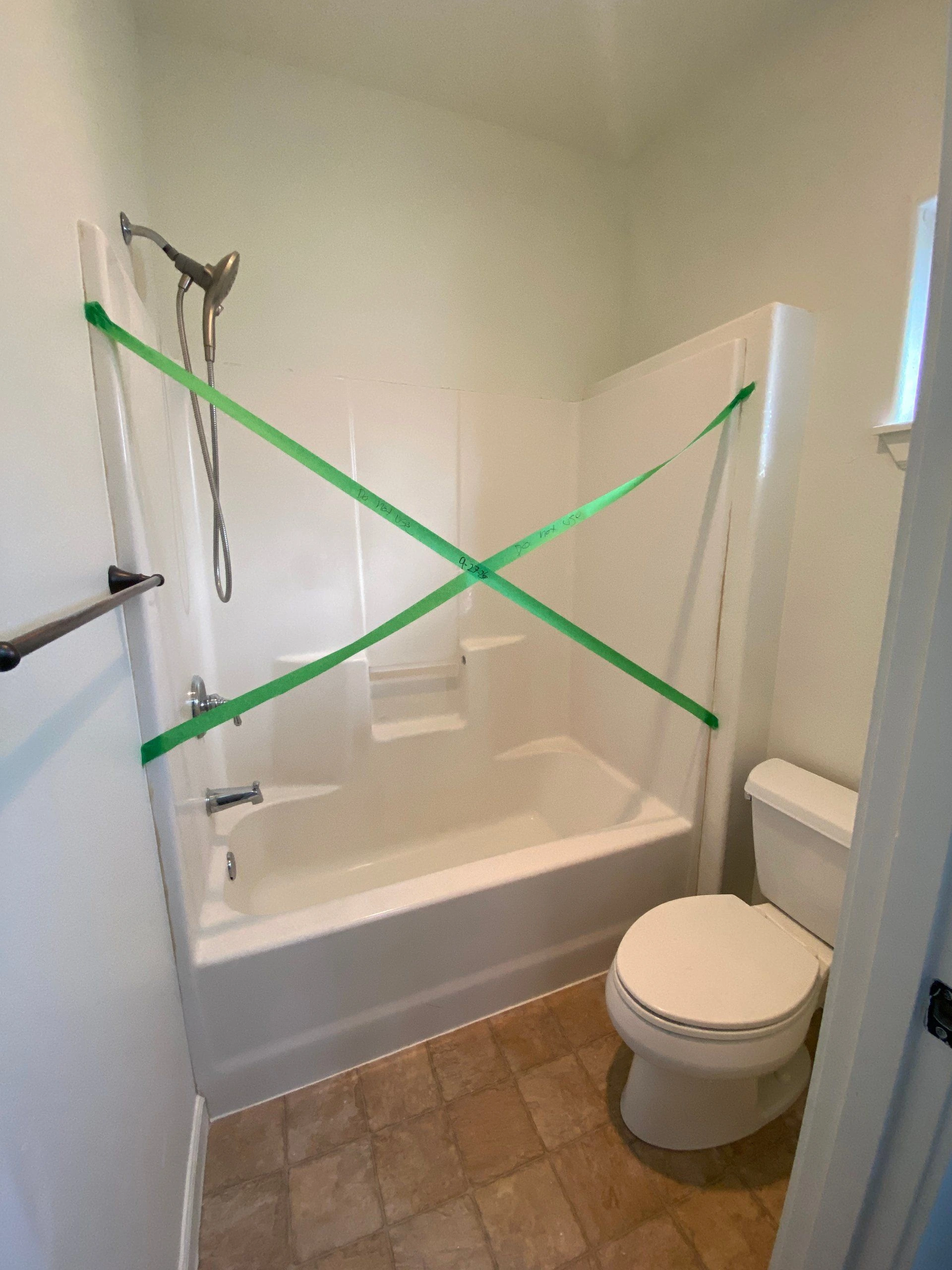 A bathtub-shower with a handheld showerhead is crossed by green tape; a toilet stands beside it on a tiled floor.