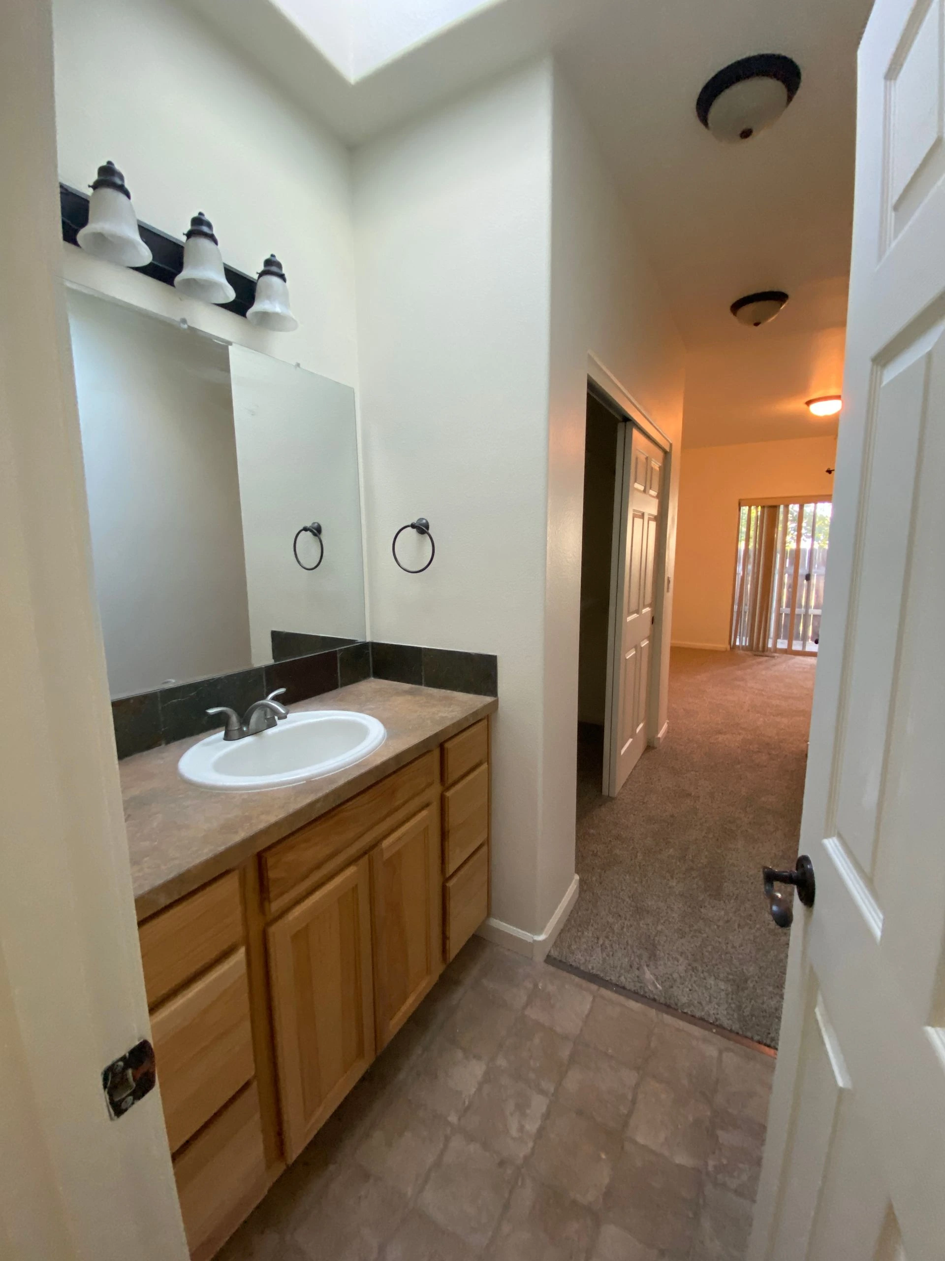 A bathroom vanity with a white sink, wood cabinets, a large mirror, and three lights opens onto a carpeted hallway.