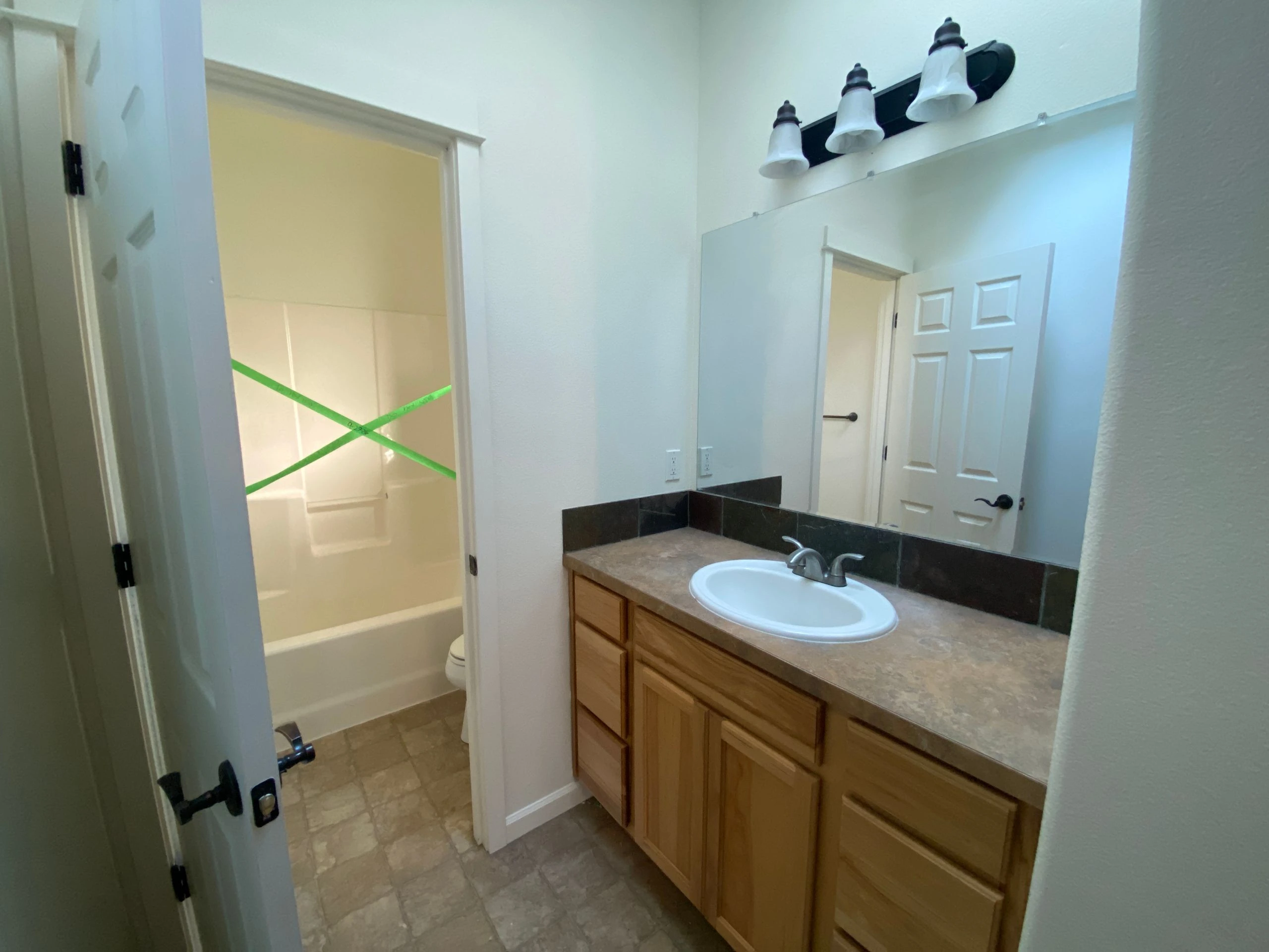 A bathroom with a wood vanity, oval sink, large mirror, and an open doorway to a tub and shower crossed by green tape.