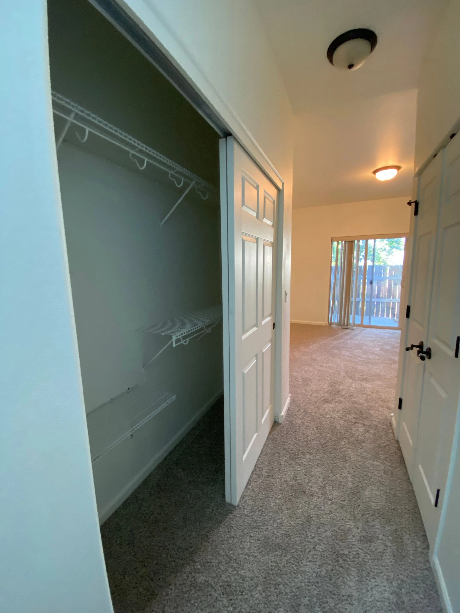 An open closet with white wire shelves and a hanging rail opens onto a carpeted room with a window and vertical blinds.
