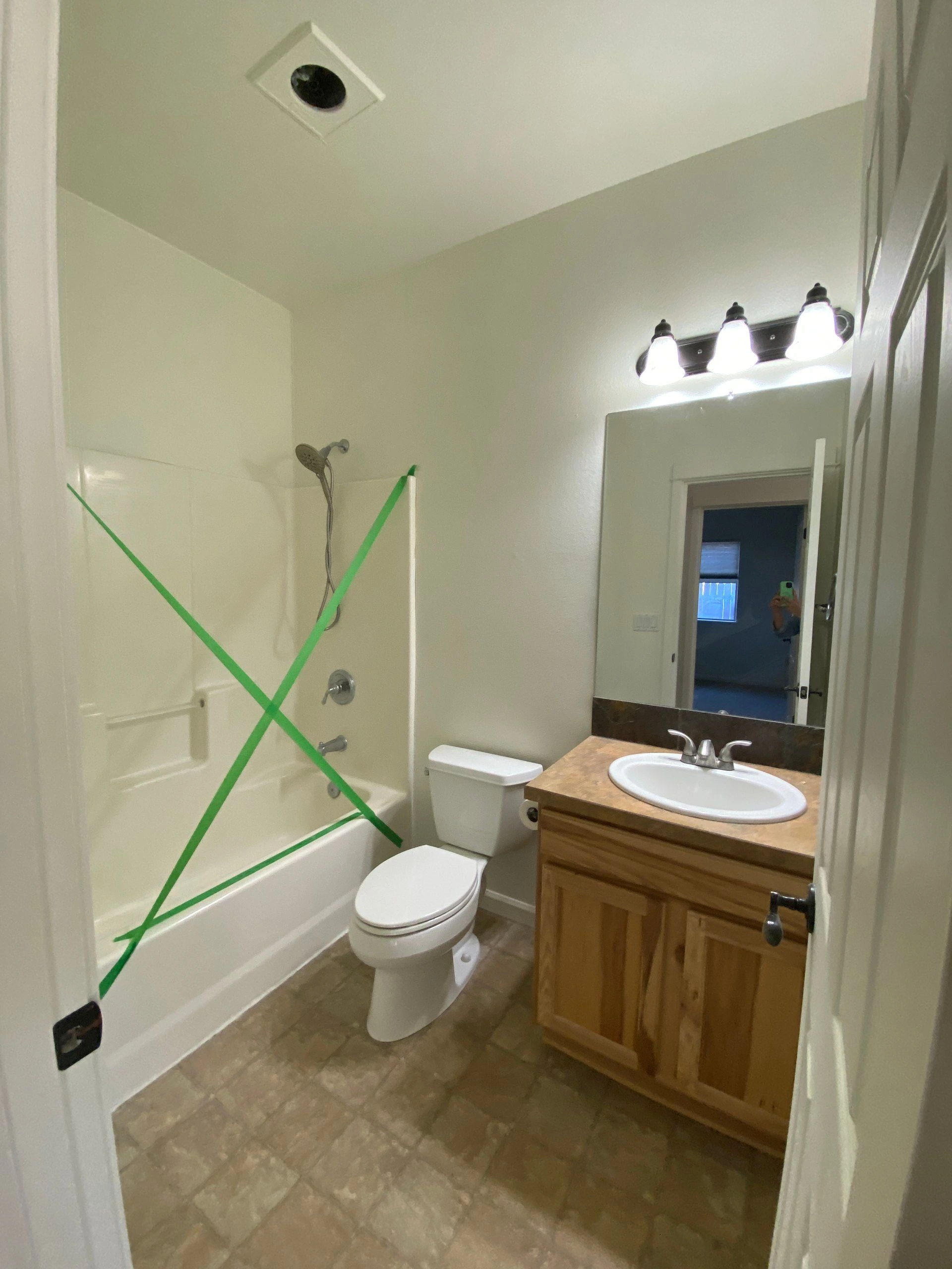 A bathroom with a bathtub marked by green tape, a toilet, and a wood vanity with a sink beneath a large mirror.
