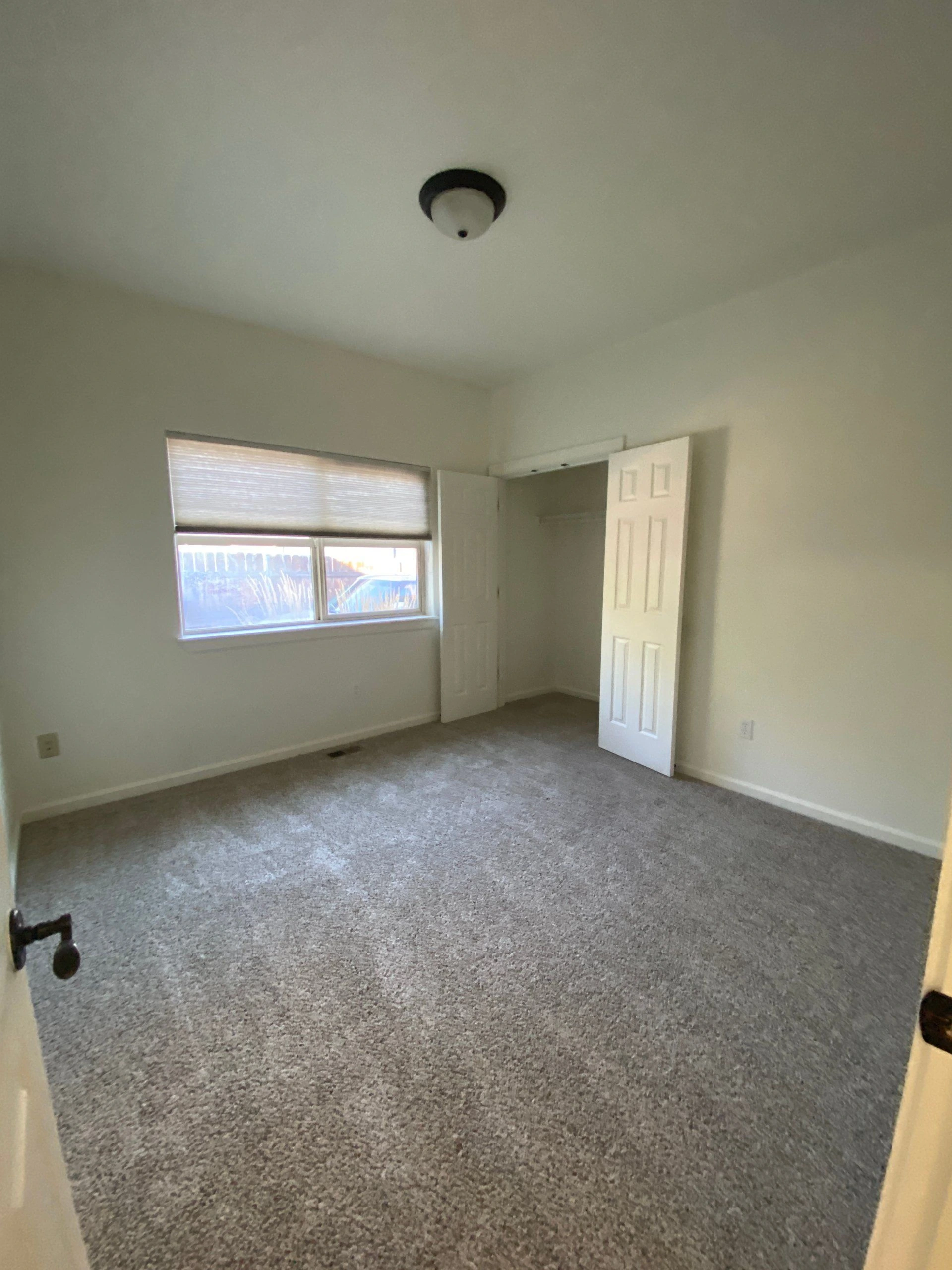 An empty room with gray carpet, a window with a lowered shade, and open closet doors.