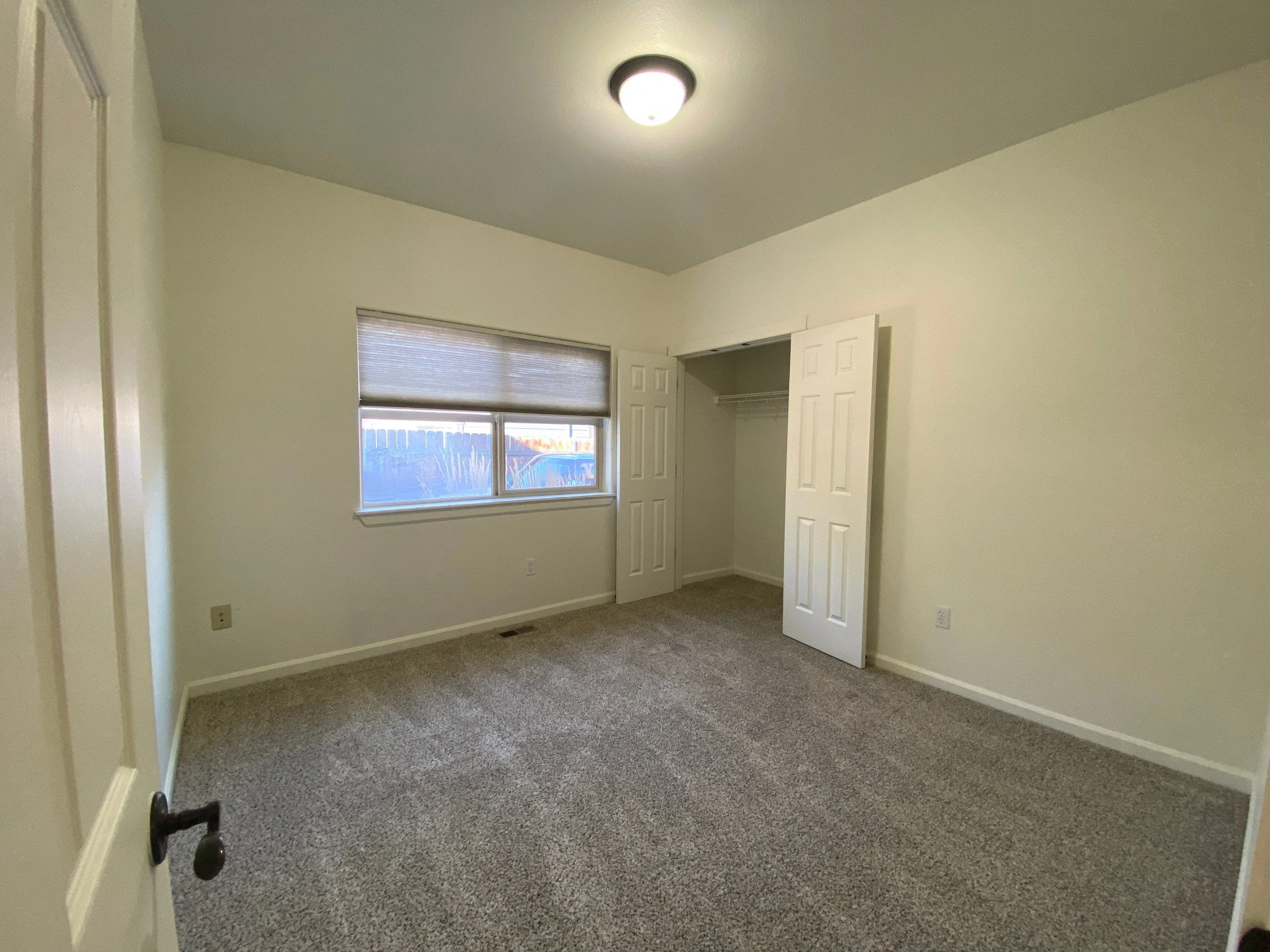 An empty carpeted room has a window with a lowered blind, an open closet, and a ceiling light.
