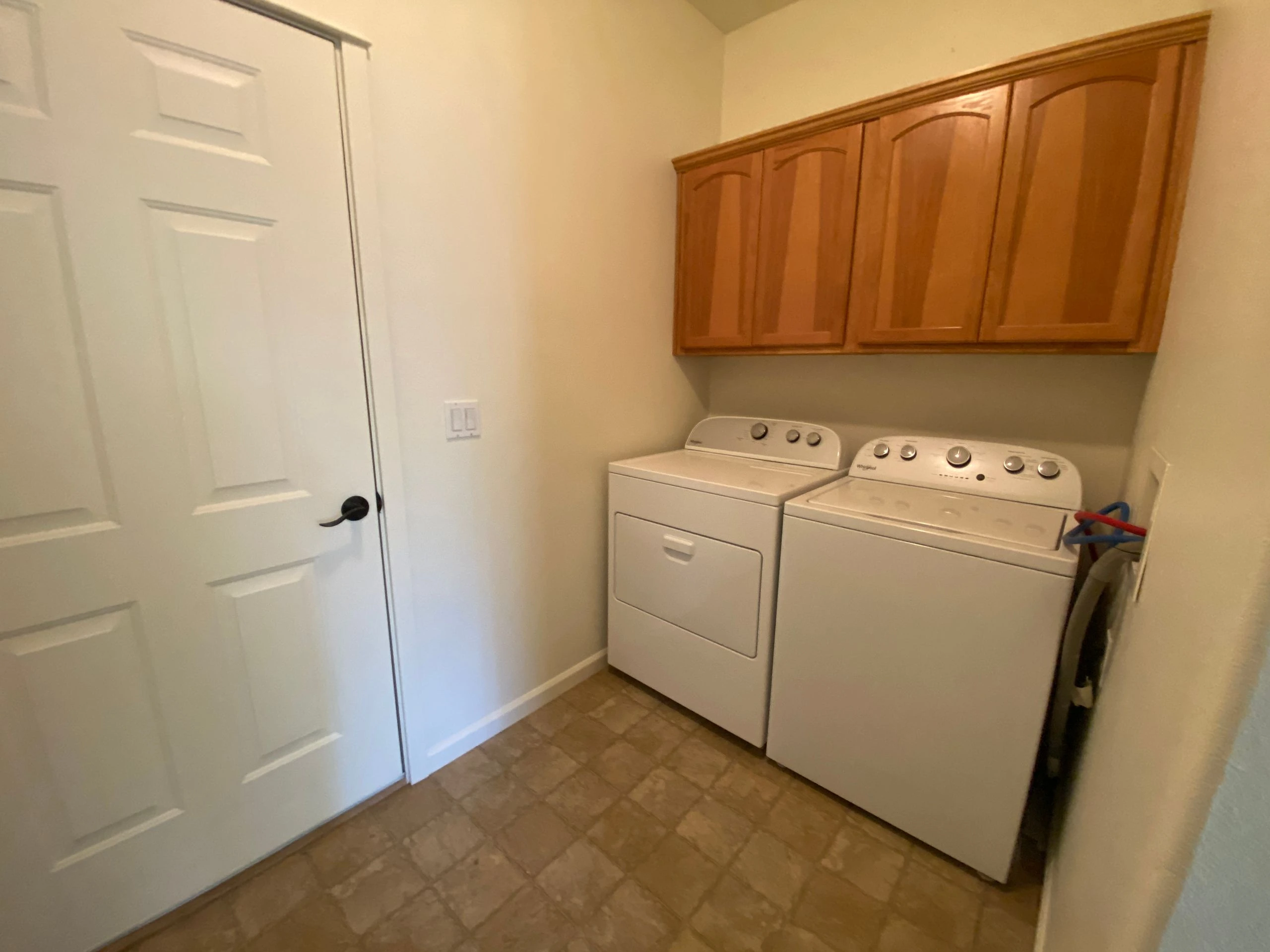 A washer and dryer sit beneath wooden cabinets in a small room with a white door and tan tile floor.