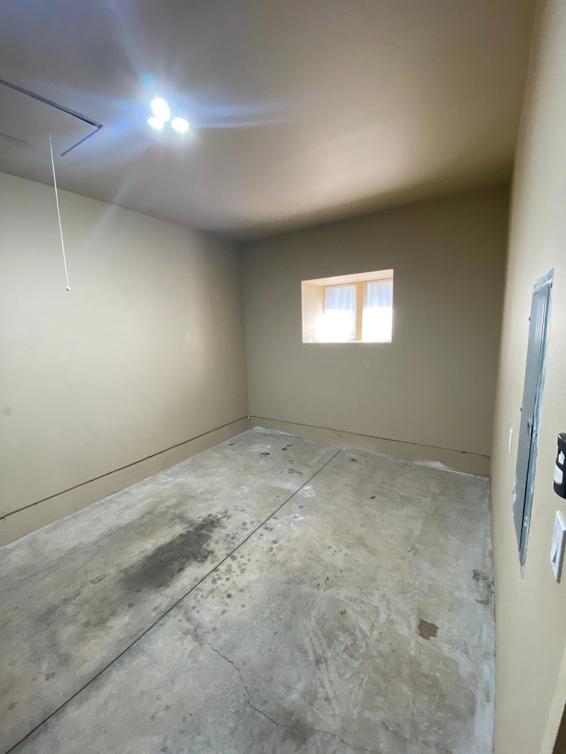 An empty room with beige walls, a small window, a lit ceiling fixture, and a stained concrete floor.