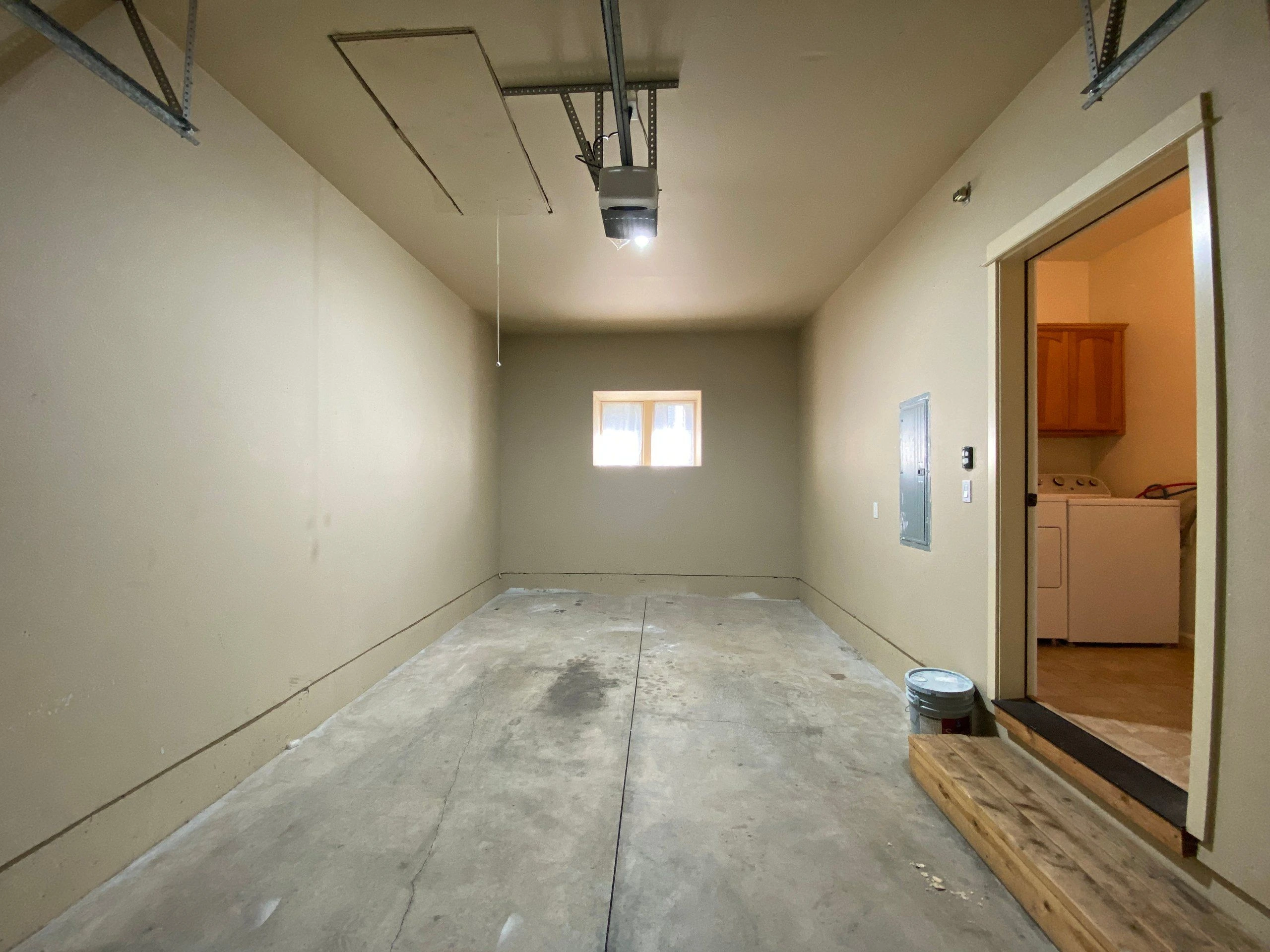 An empty garage with a concrete floor, ceiling-mounted door tracks, and a small window on the far wall; laundry machines are visible through an open doorway.