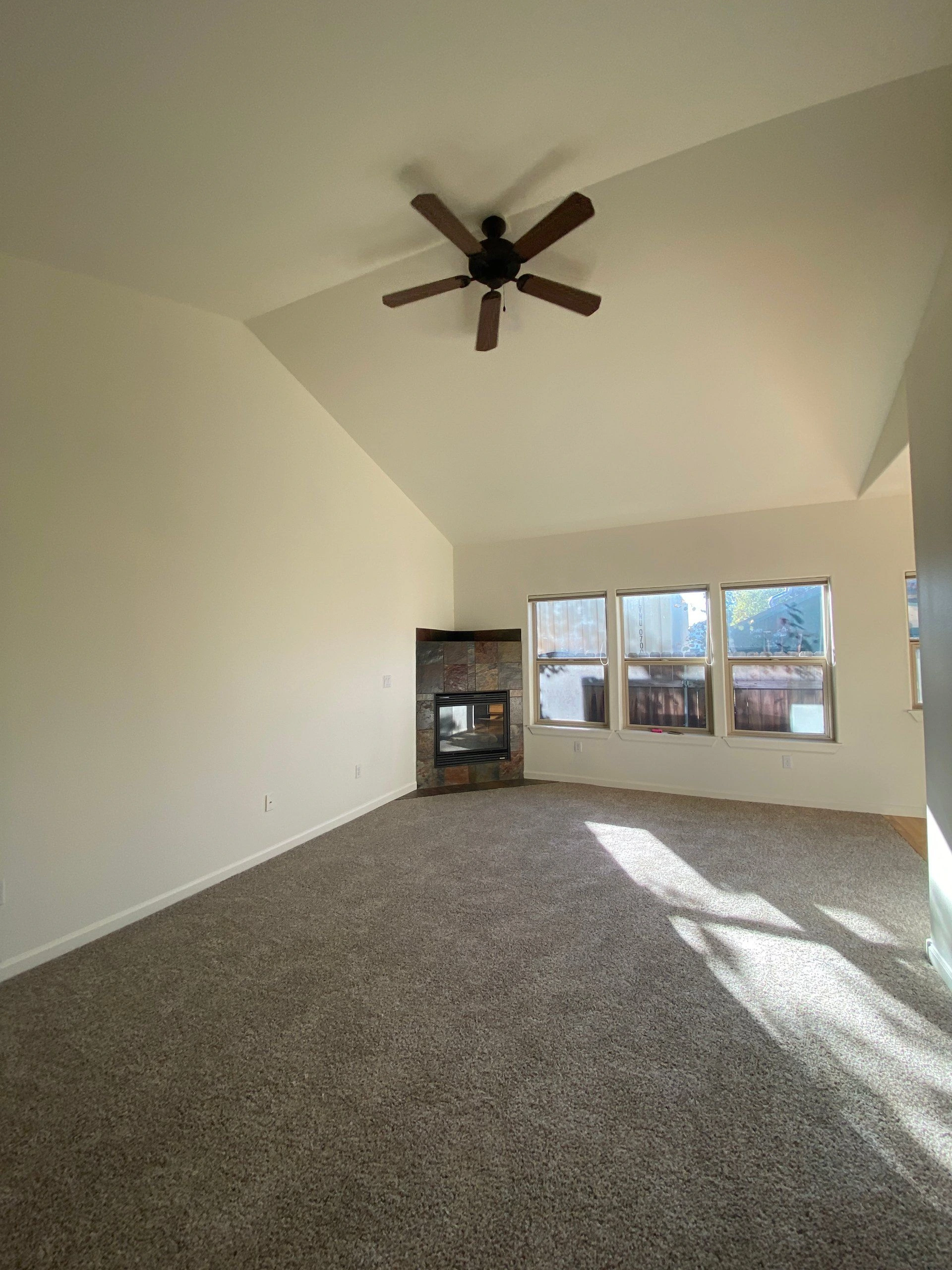 An empty carpeted room has a vaulted ceiling, a five-blade ceiling fan, a tiled fireplace, and several windows.