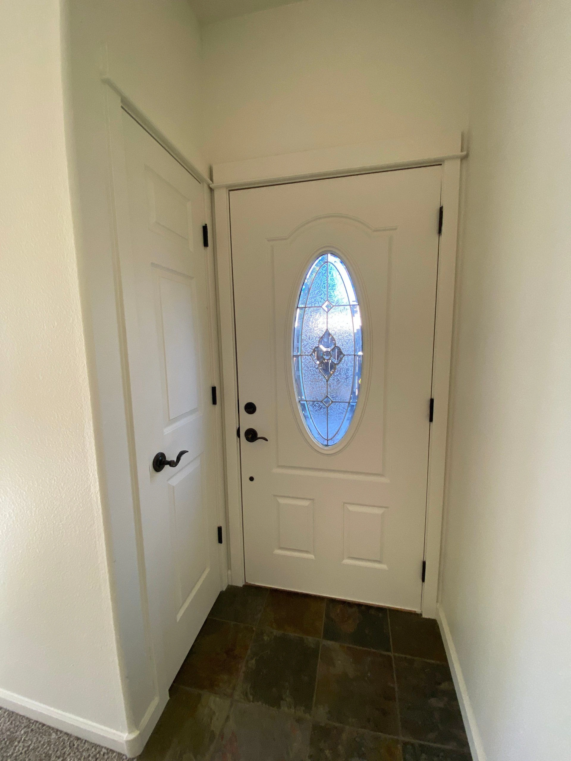 A white front door with an oval decorative glass panel stands above a dark tiled floor, beside a partly open white door.