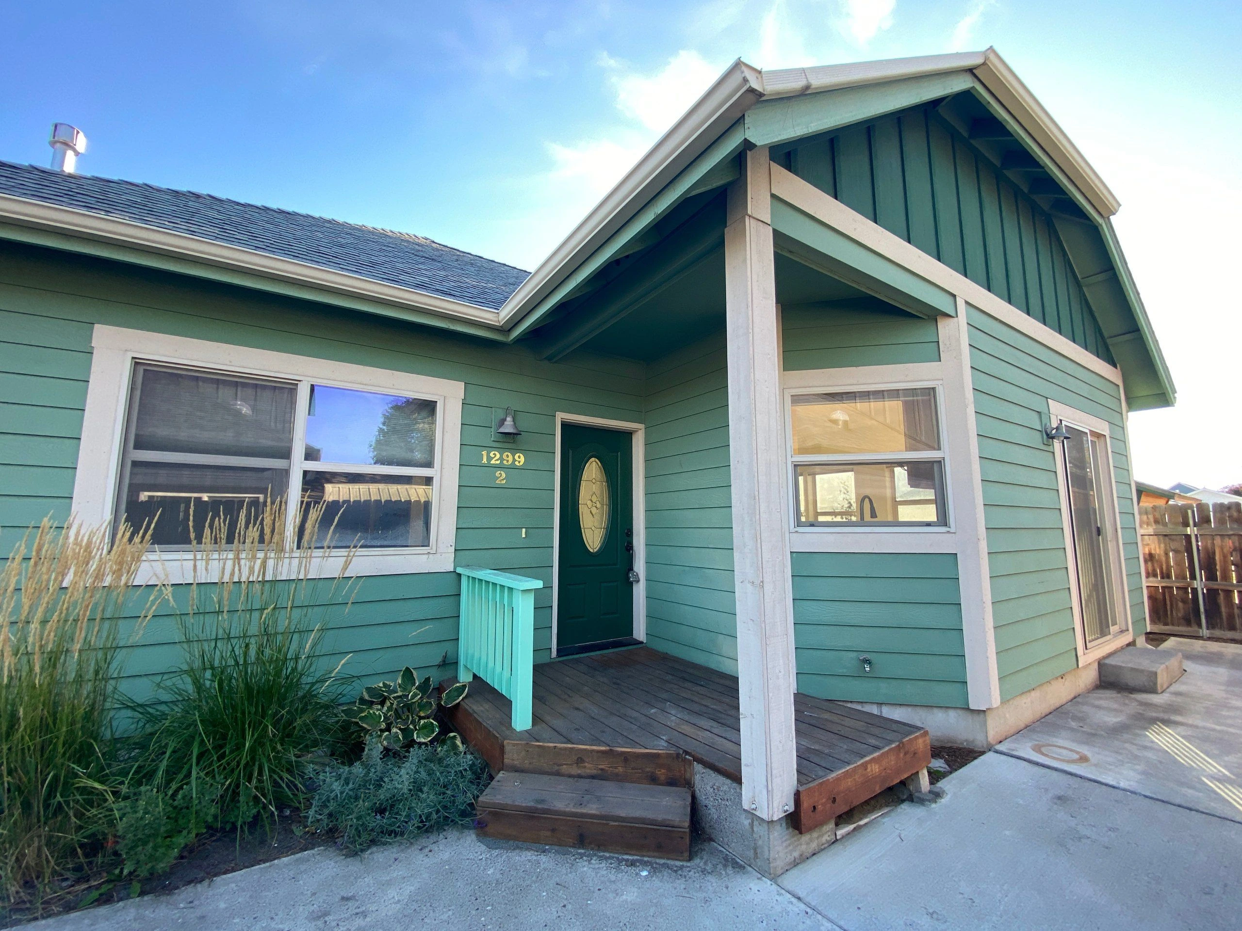 A teal-sided house has white trim, a gabled roof, and a small covered wooden porch with a dark front door and windows.