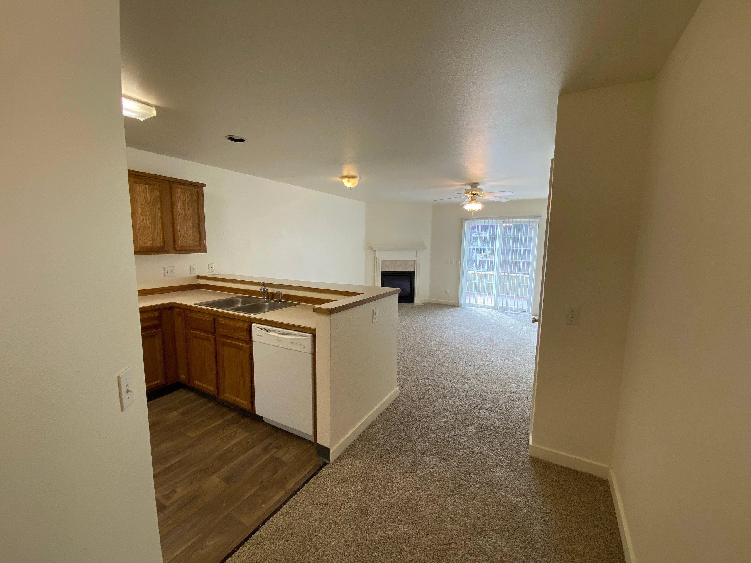 A kitchen with wood cabinets, a double sink, and dishwasher opens onto a carpeted room with a fireplace, ceiling fan, and sliding glass door.