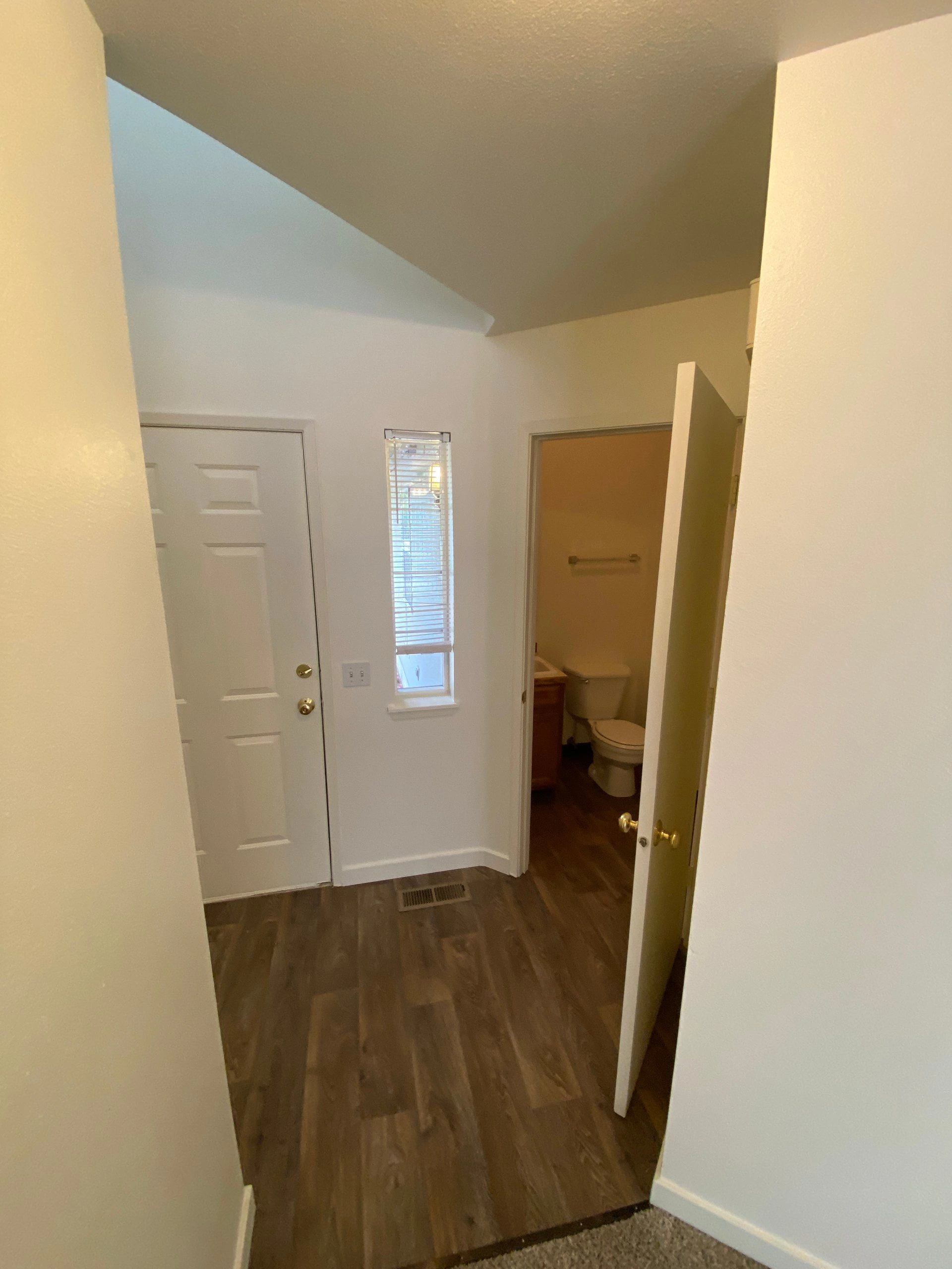 A hallway with dark wood flooring, a narrow window with blinds, a closed door, and an open bathroom doorway.