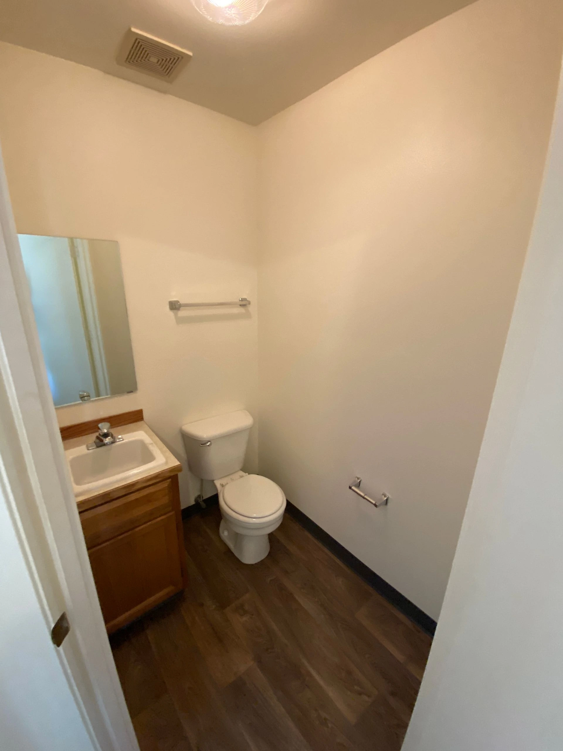 A small bathroom has a toilet, a sink with a wooden vanity, a mirror, towel bars, and dark wood-look flooring.