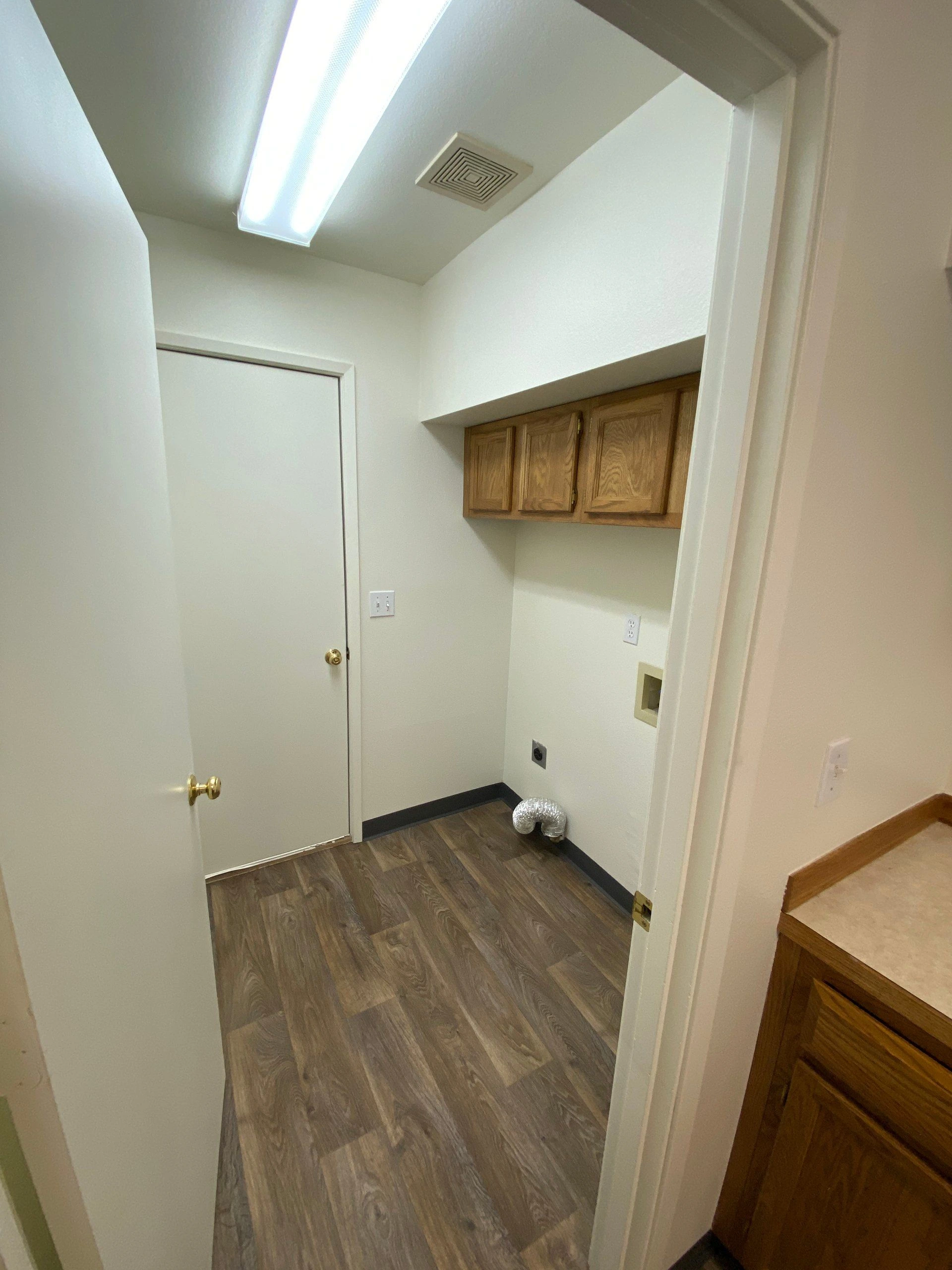A narrow room has brown plank flooring, wooden wall cabinets, a closed white door, and a ceiling light and vent.
