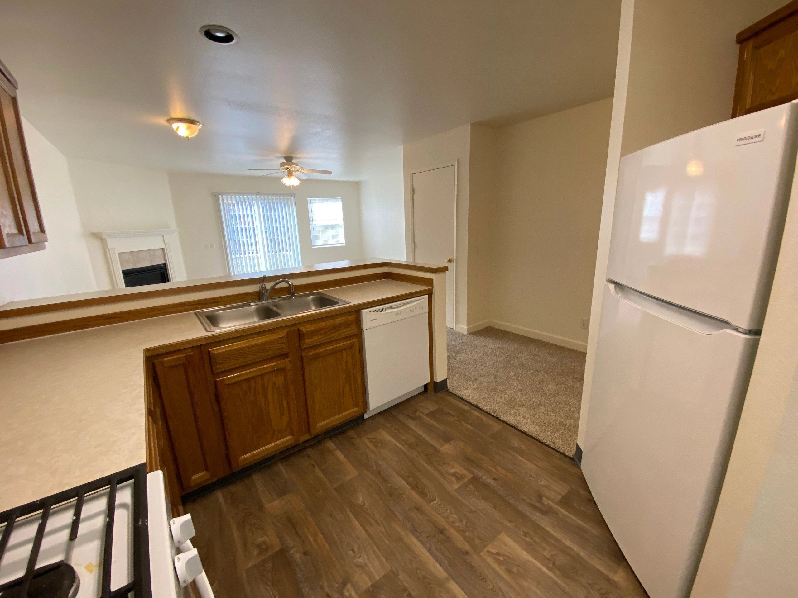 A kitchen with wood cabinets, a double-basin sink, dishwasher, refrigerator, and a counter opening onto a room with a fireplace and windows.