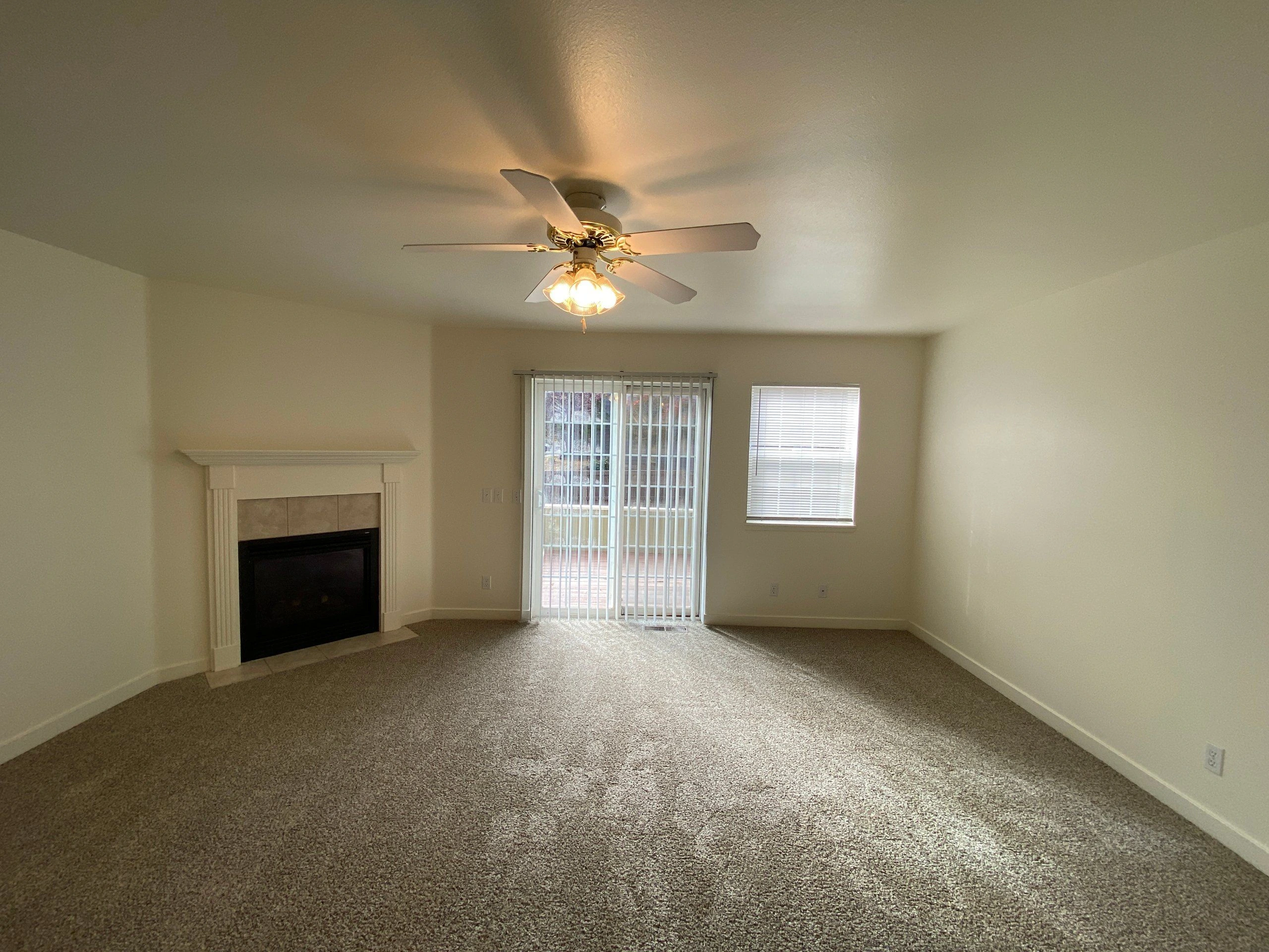An empty carpeted room with a fireplace, ceiling fan with lights, sliding glass door with vertical blinds, and a window with horizontal blinds.