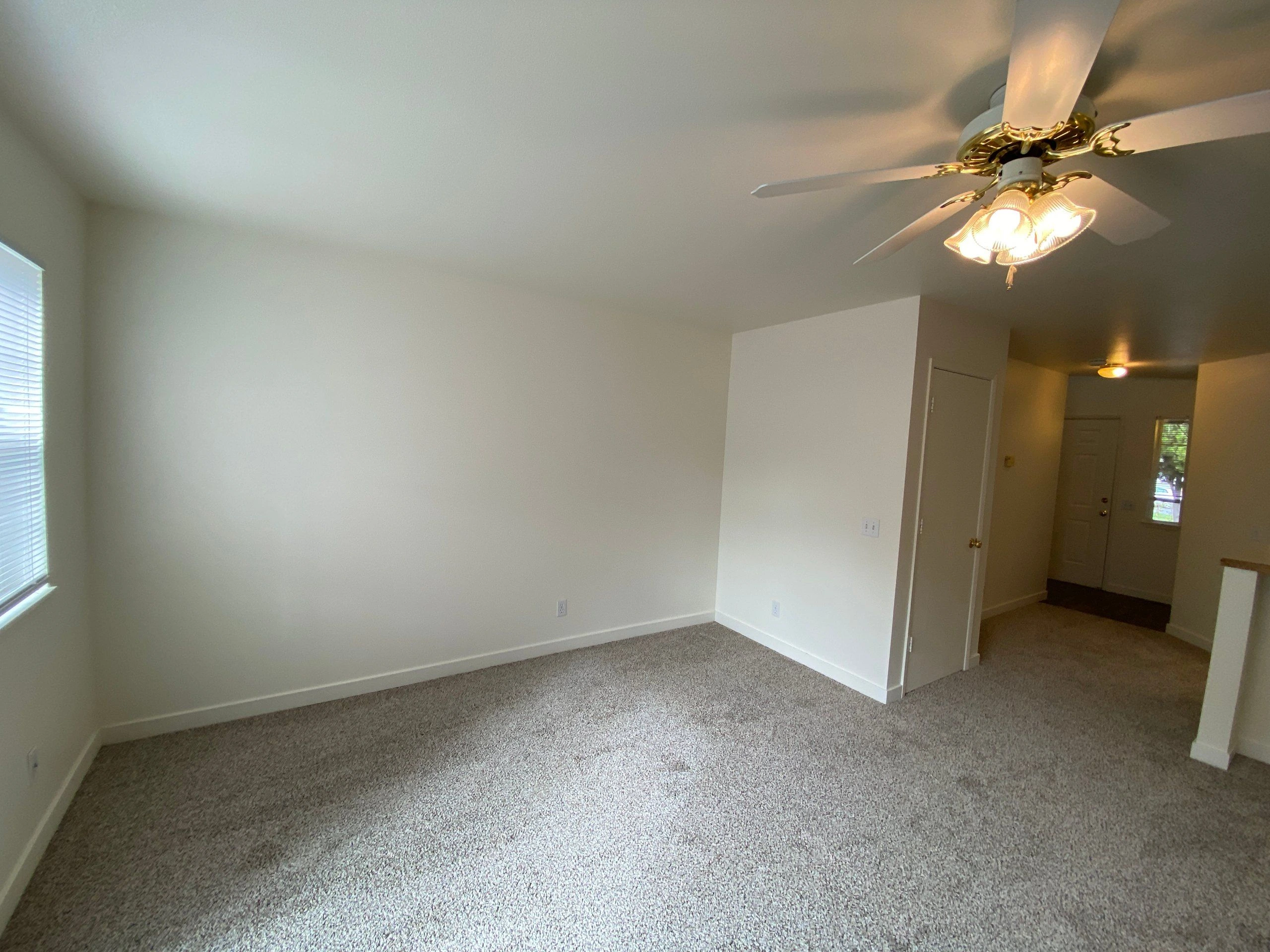 An empty carpeted room has a window with blinds, a ceiling fan with lights, and an open doorway to a hall.