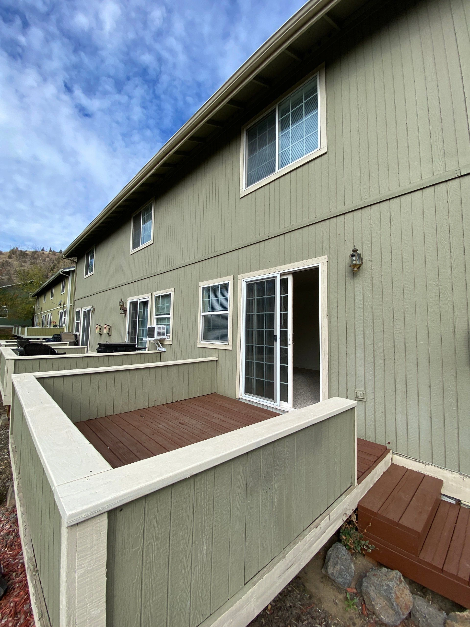 A raised wooden deck with pale green railings sits beside a green-sided building with windows and an open sliding glass door.