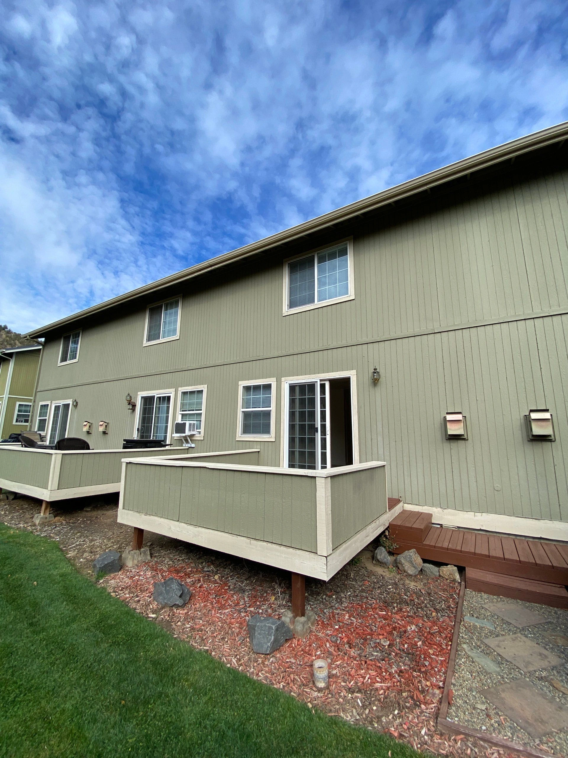 A green-sided building with several windows and an open sliding door faces a raised, railed deck beneath a cloudy blue sky.