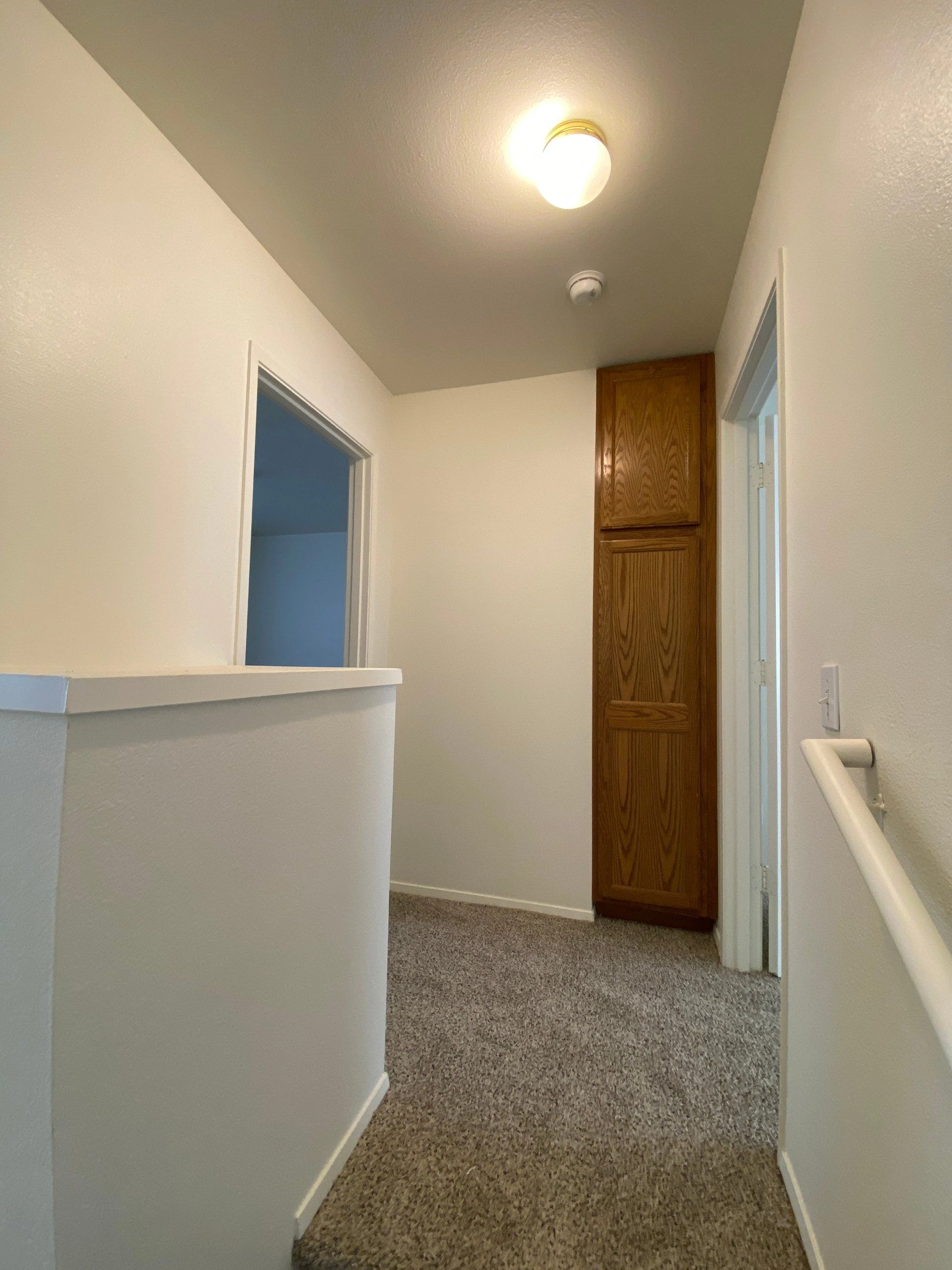 A carpeted hallway with white walls, a lit ceiling fixture, open doorways, and a tall wooden cabinet.